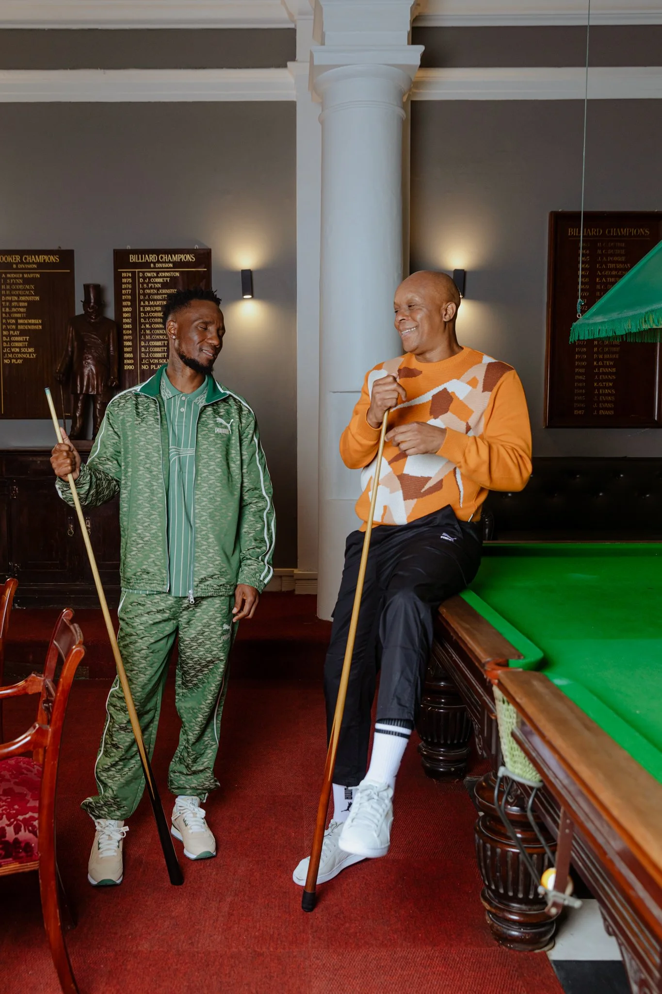 Two men talking near a pool table in a room with plaques on the wall and a statue. The man on the left wears a green tracksuit and holds a pool cue. The man on the right, sitting on the pool table's edge, wears an orange patterned sweater, black pant