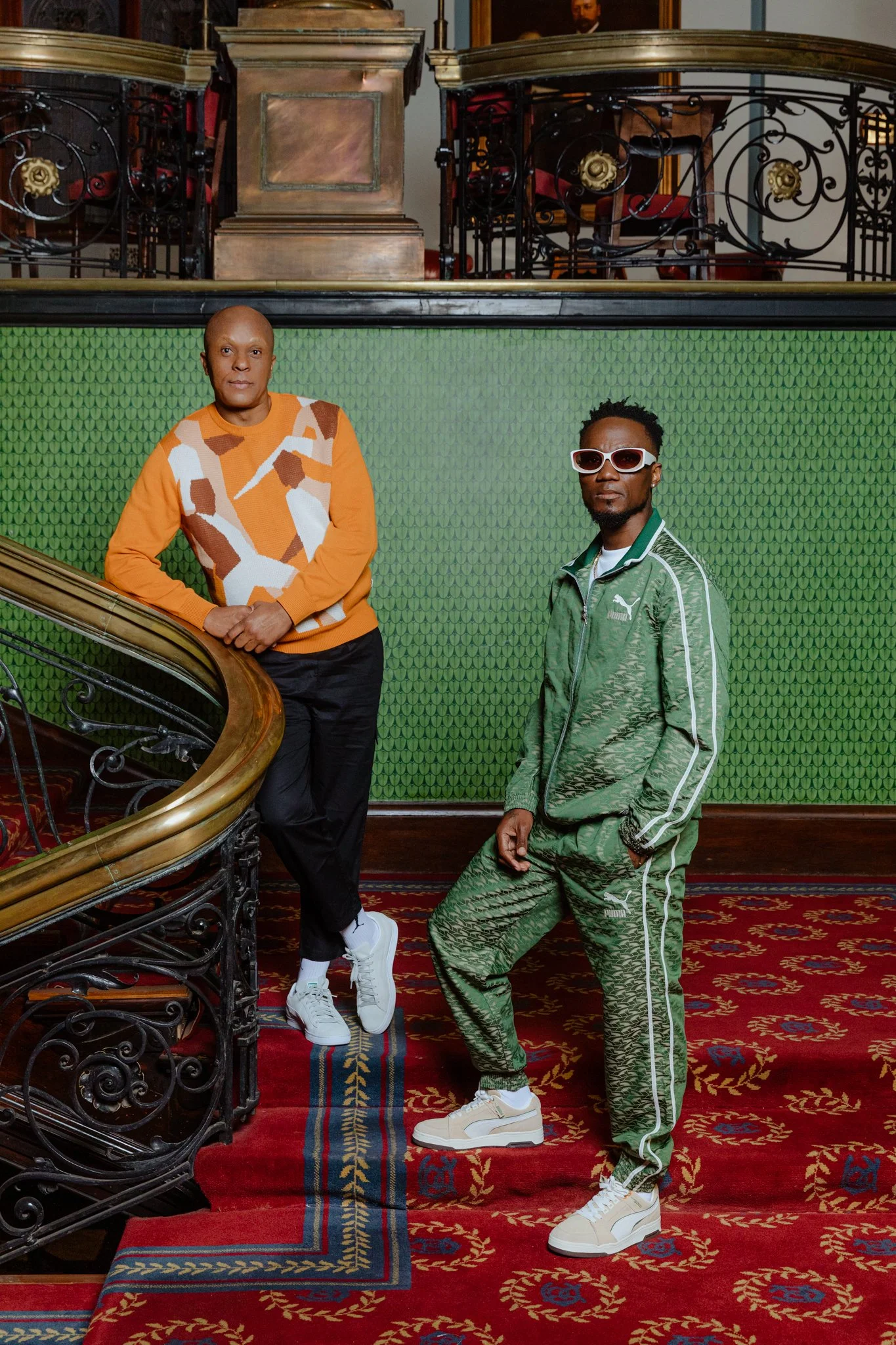 Two men standing on a red patterned carpet in front of a green textured wall inside a building with ornate black iron railing and a balcony above. The man on the left is leaning against a staircase railing, wearing a colorful sweater, black pants, an