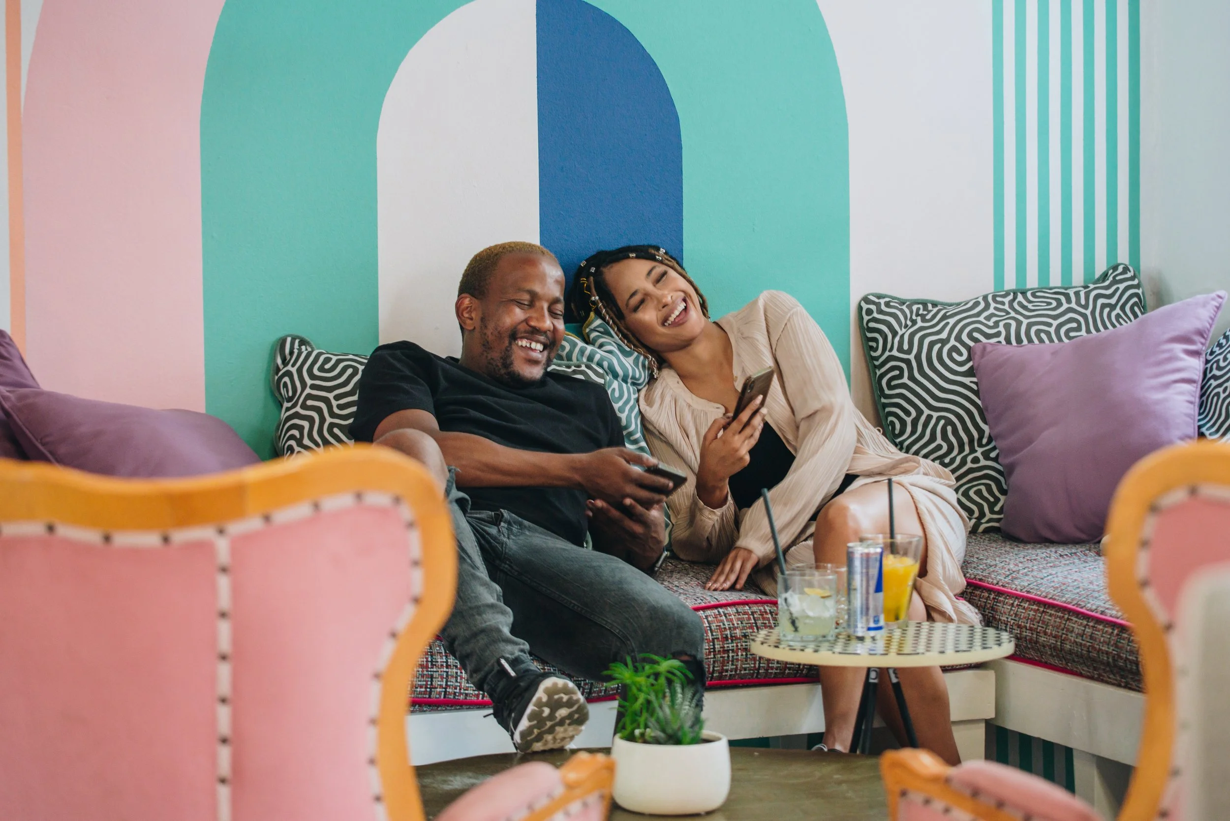 Two people sitting on a colorful couch, smiling and looking at their phones. They are surrounded by glasses of drinks and pillows, in a room with a vibrant, pastel-colored wall.