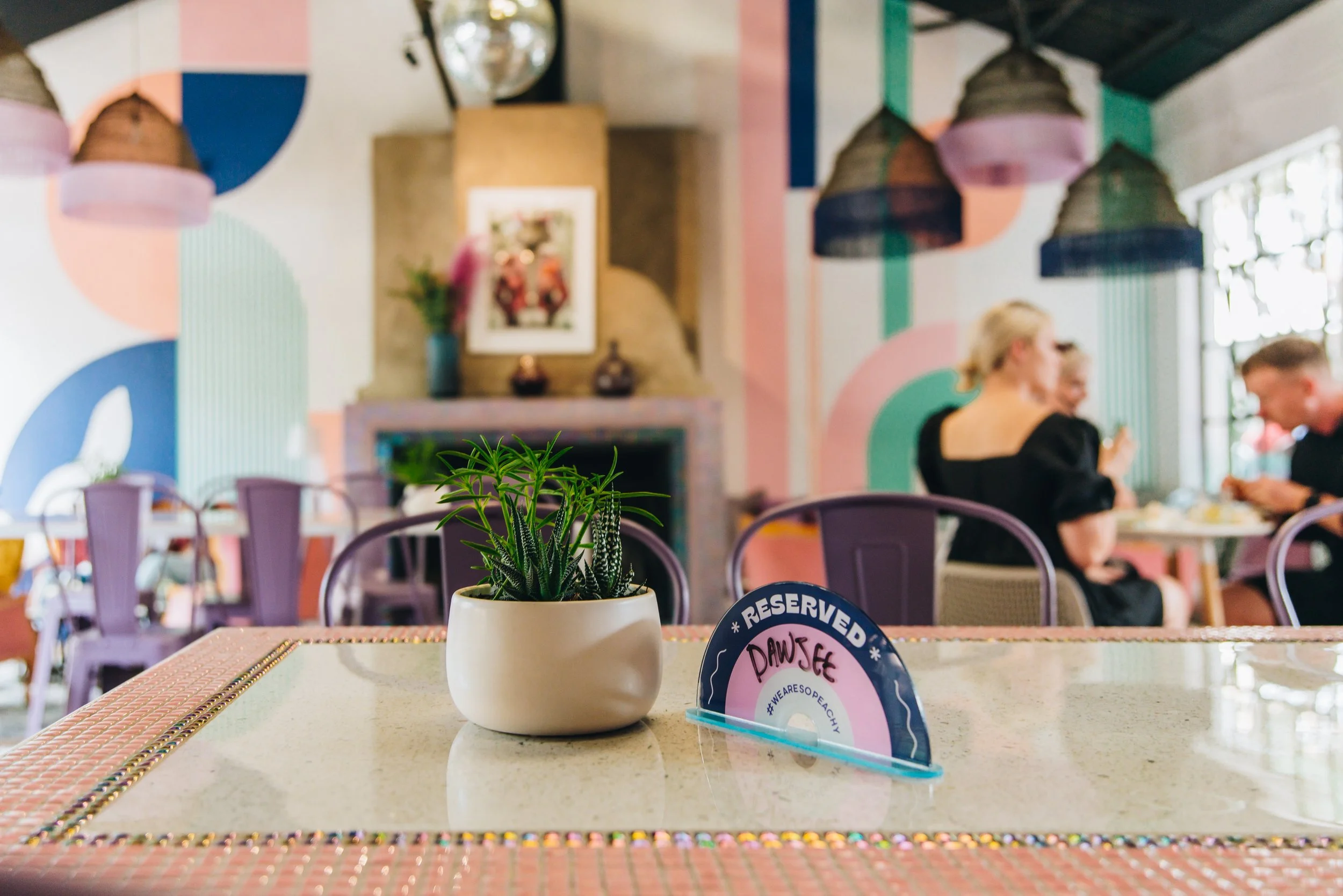Indoor restaurant or café with a plant in a white pot on the foreground table, reserved sign with the name 'Dawsee', blurred background of people dining, colorful geometric and striped wall design, hanging lamps, and a fireplace with decorations.