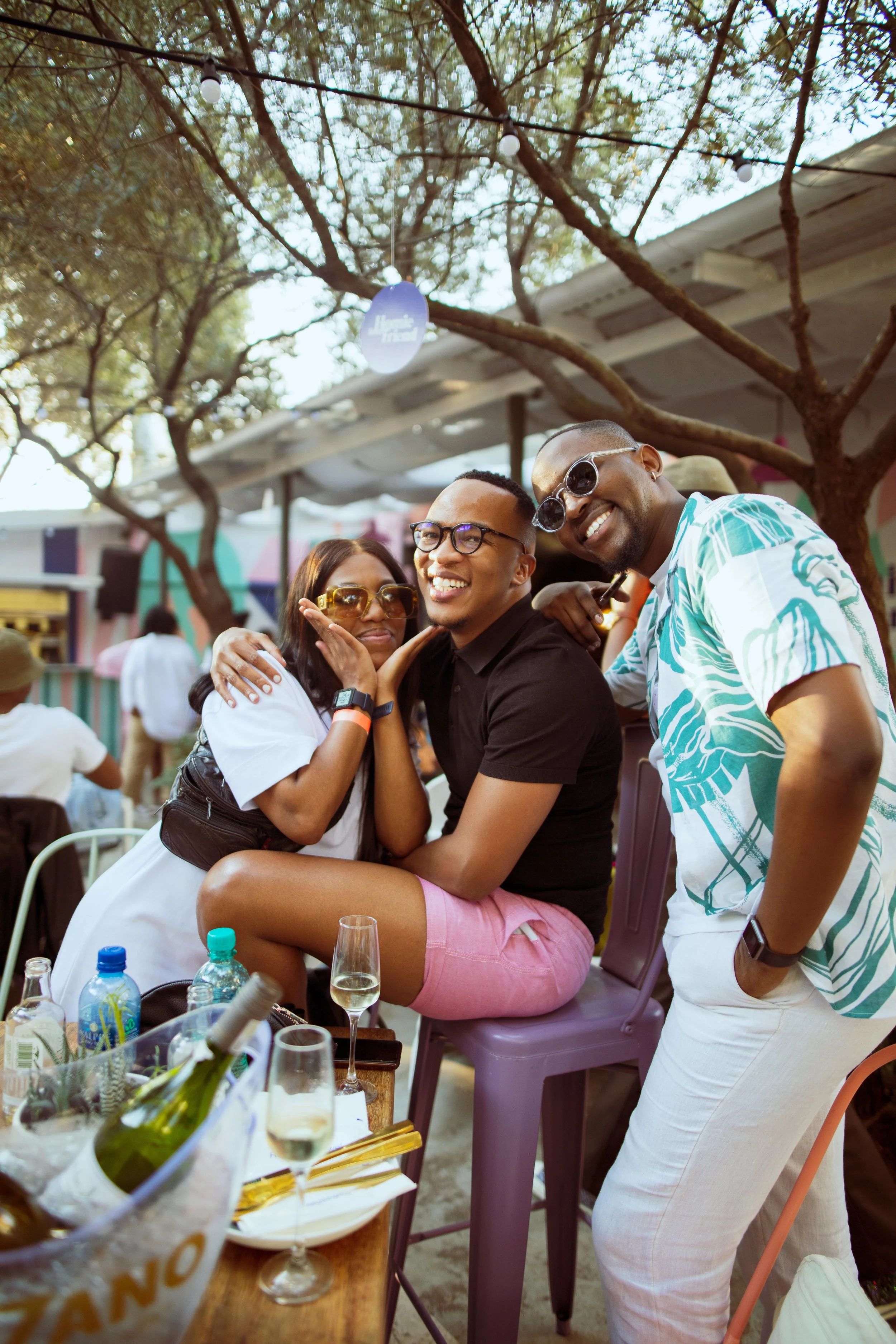 Three people smiling and enjoying a moment together at an outdoor gathering, with one seated on a purple stool and two standing beside her, under string lights and trees.