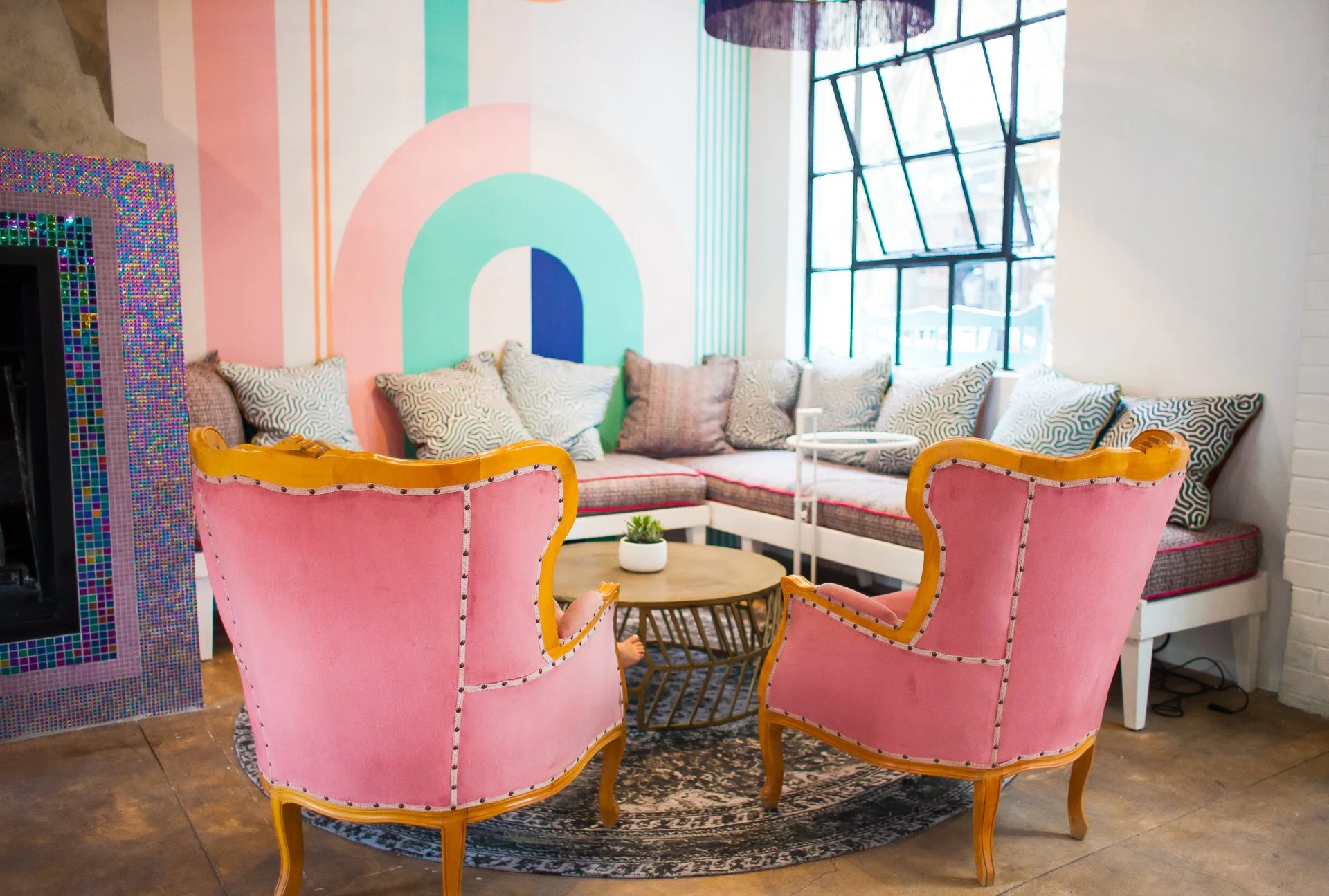 Cozy sitting area with vintage pink armchairs facing a round wooden coffee table, a pink and grey patterned sectional sofa with multiple pillows, and a colorful wall mural with geometric shapes, all by a large window with grid panes.