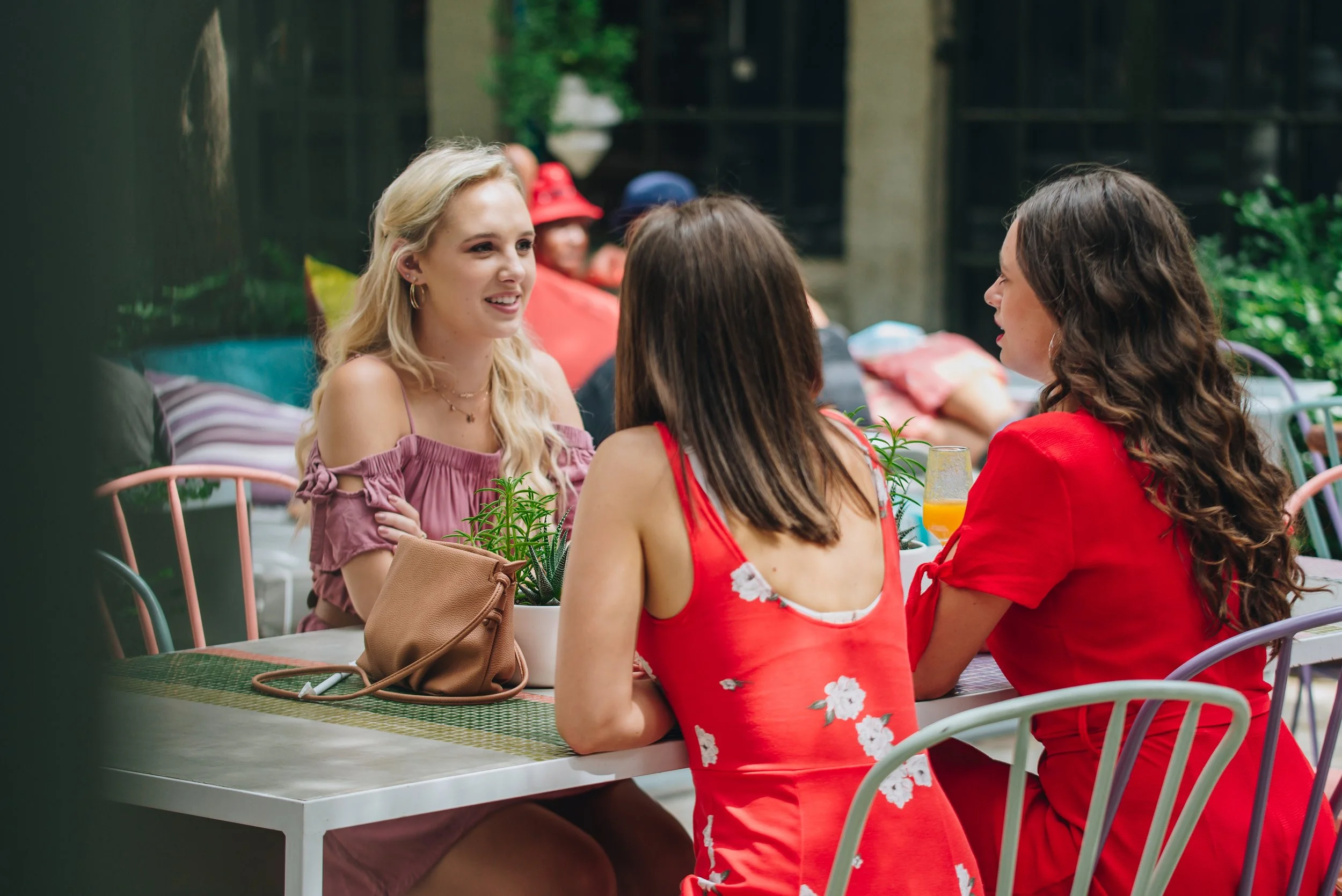 Three women sitting at a table outdoors, engaged in conversation, with drinks and a handbag on the table, surrounded by colorful chairs and lush greenery.