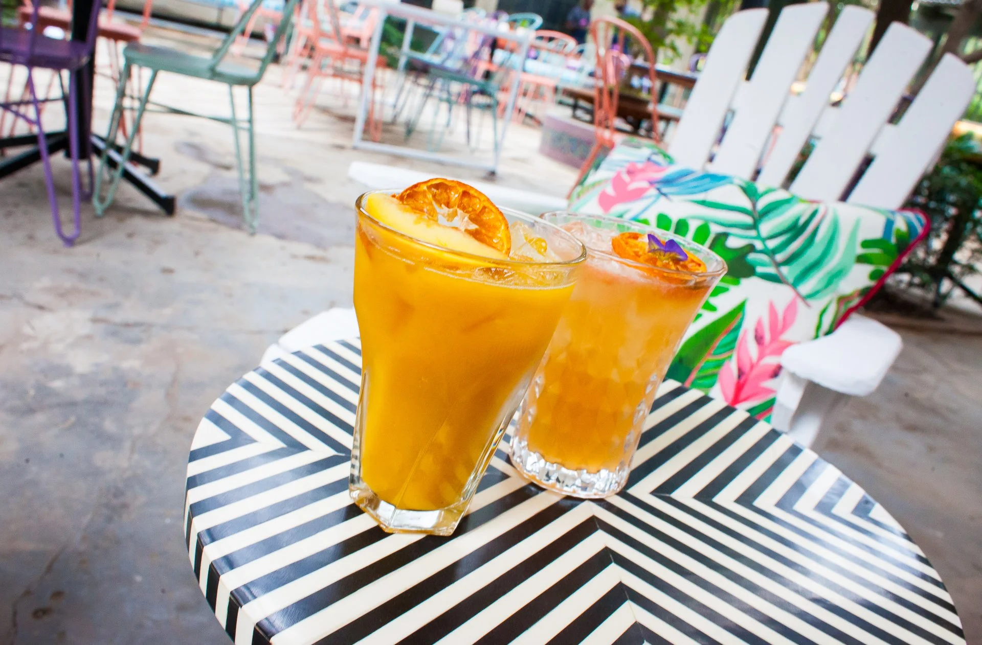 Two tropical cocktails or drinks with orange slices on a black and white striped table in an outdoor setting.