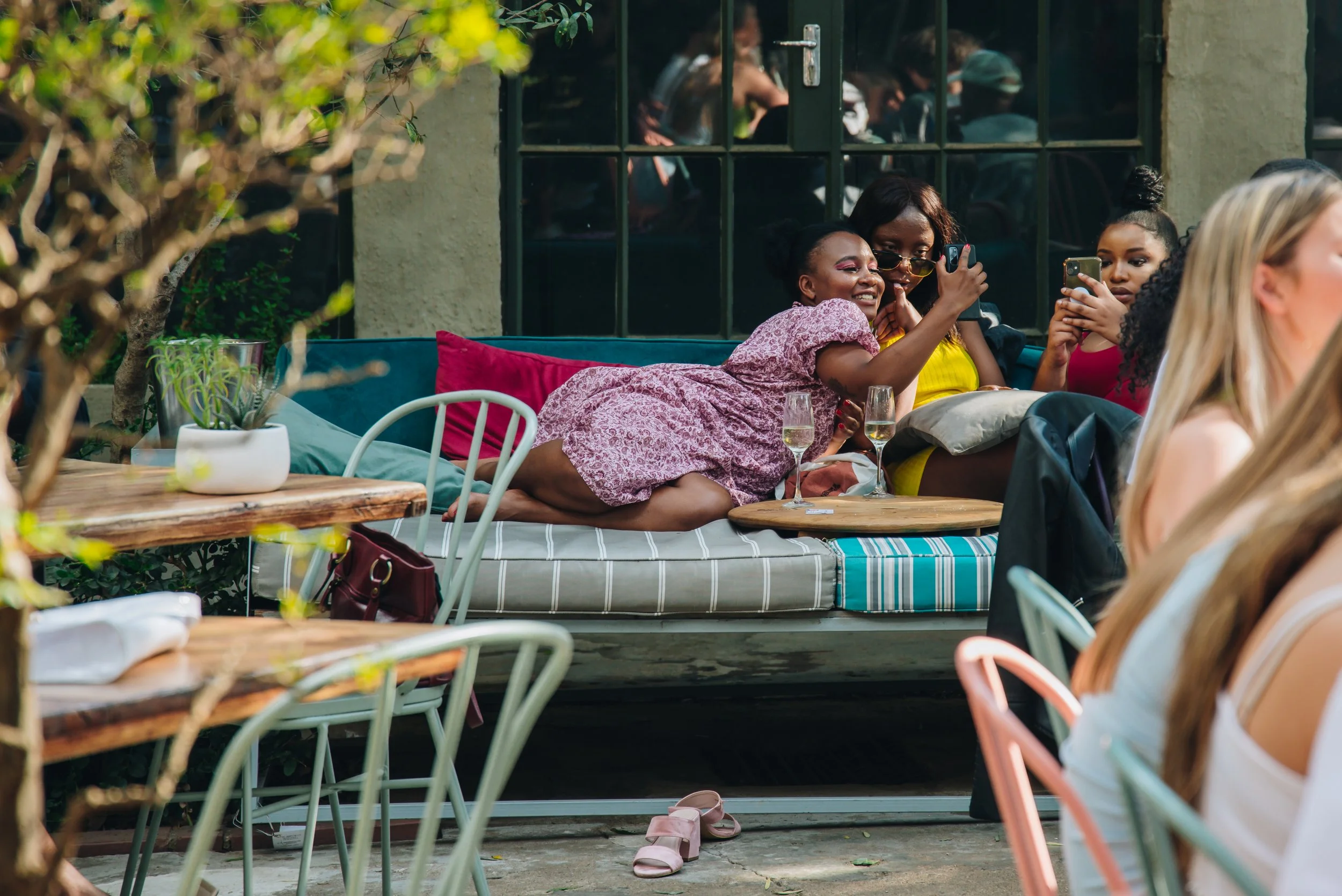 Three women on an outdoor patio, taking selfies and smiling, surrounded by tables, chairs, and plants.