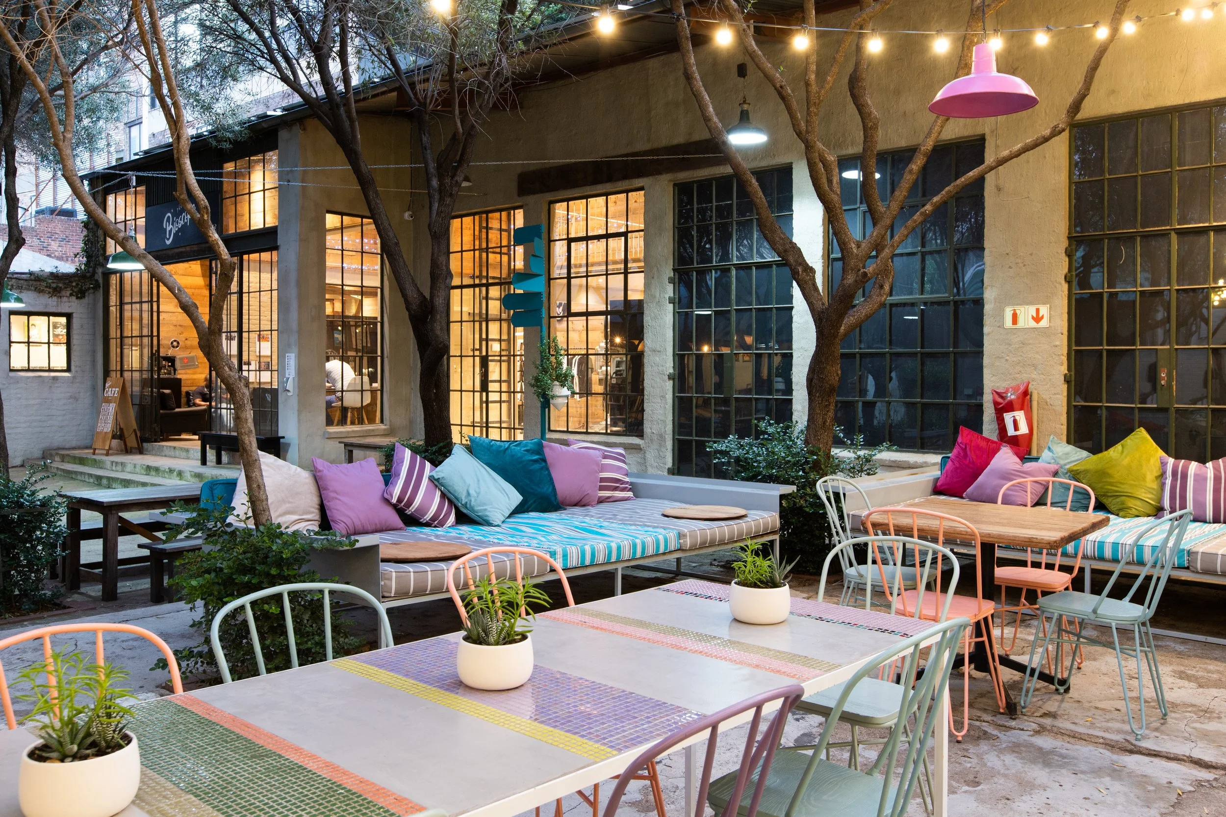 Outdoor patio of a cafe or restaurant with colorful chairs and cushions, potted plants, and string lights, featuring a building with large glass windows in the background.