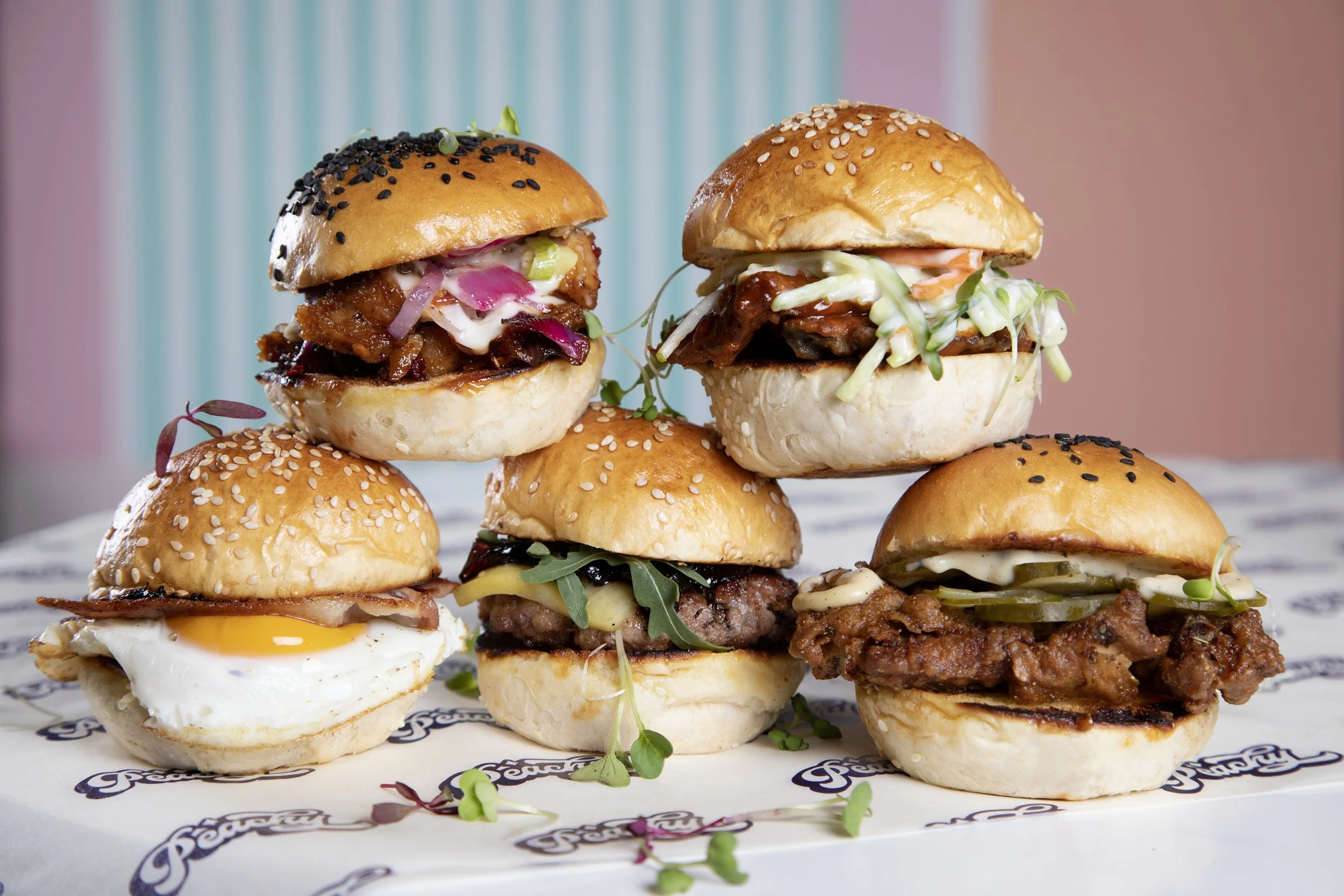 Six mini burgers with various toppings, including an egg, fried chicken, beef patty, and veggie, arranged on white paper with a patterned background.