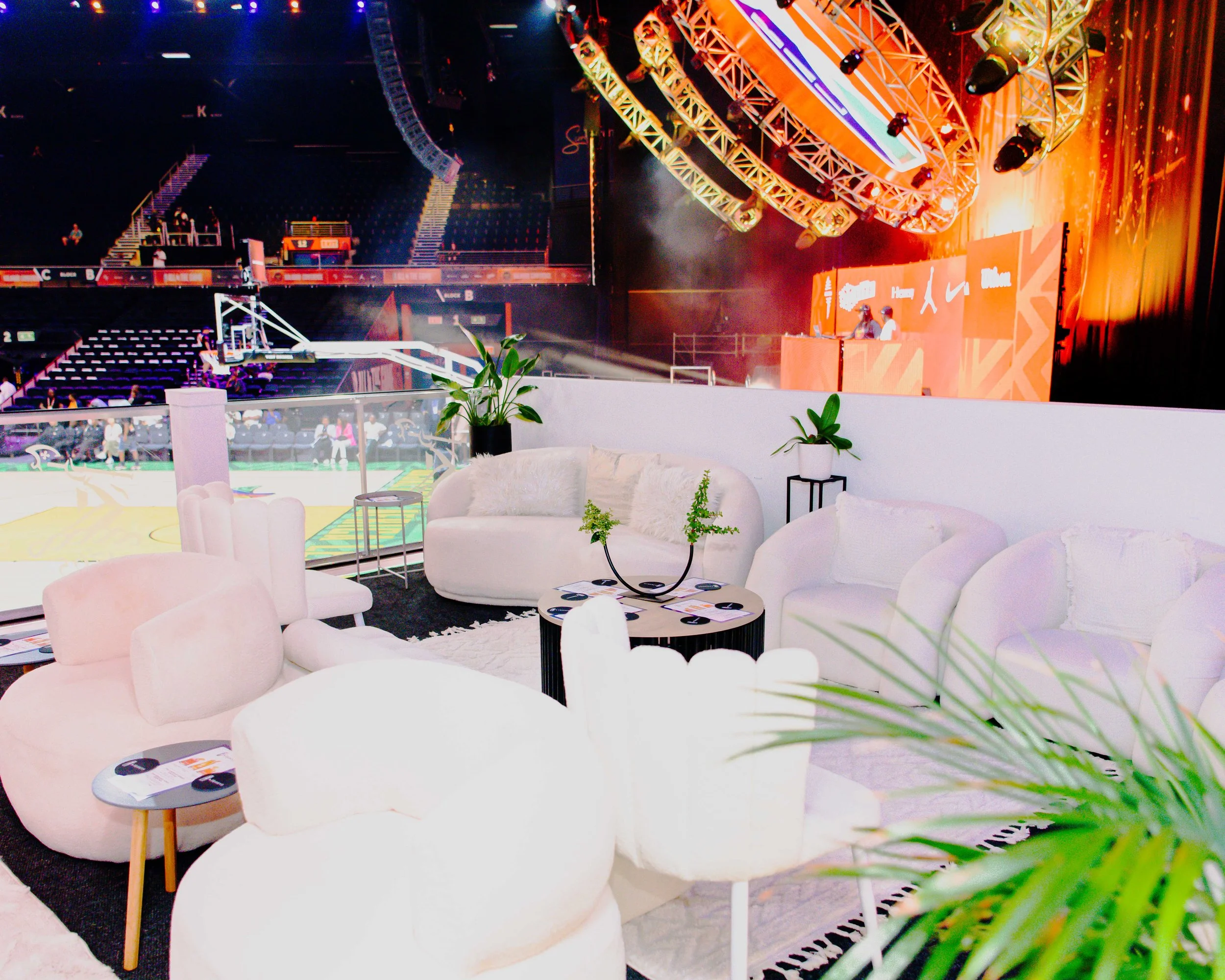 A lounge area with white sofas and chairs, decorated with green plants, overlooking a large, brightly lit basketball arena with a game in progress on the court.