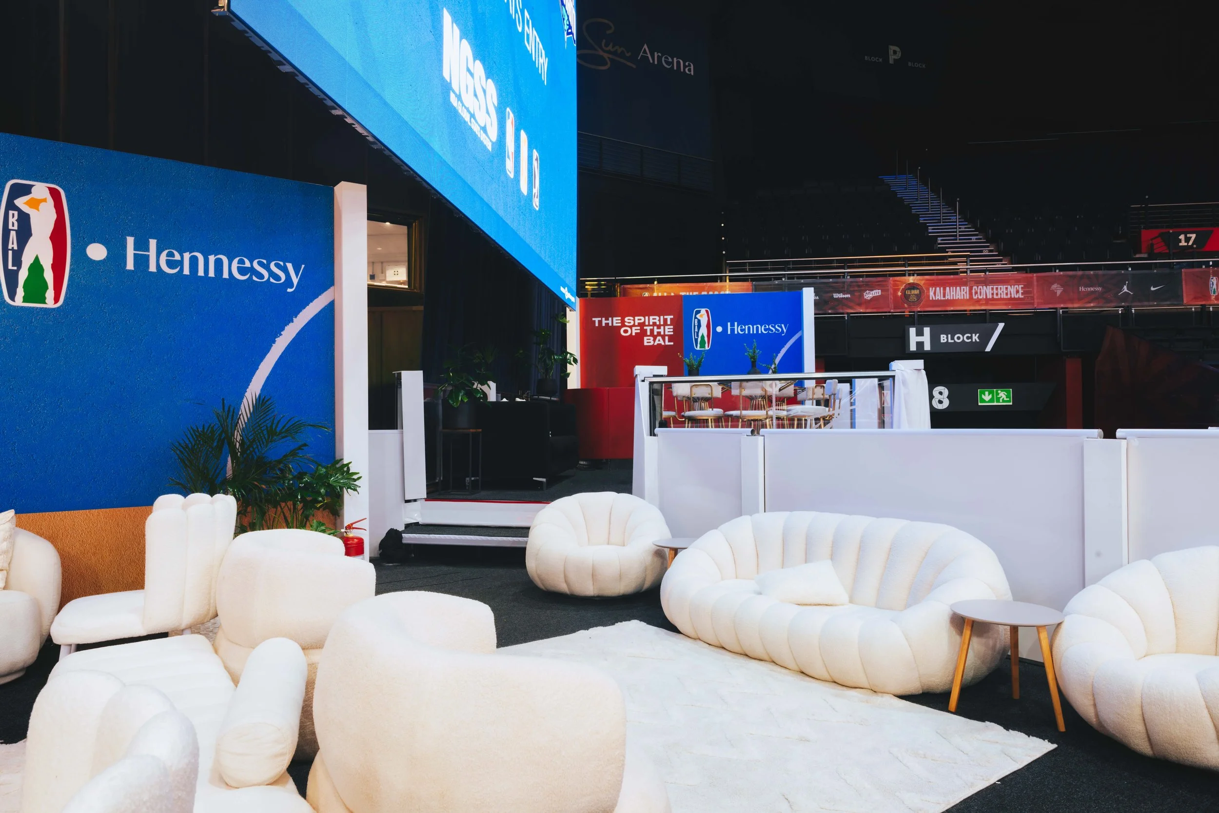 Empty seating area with cream-colored, plush chairs and small tables in front of large blue, red, and white banners displaying NBA and Hennessy logos inside a sports arena.