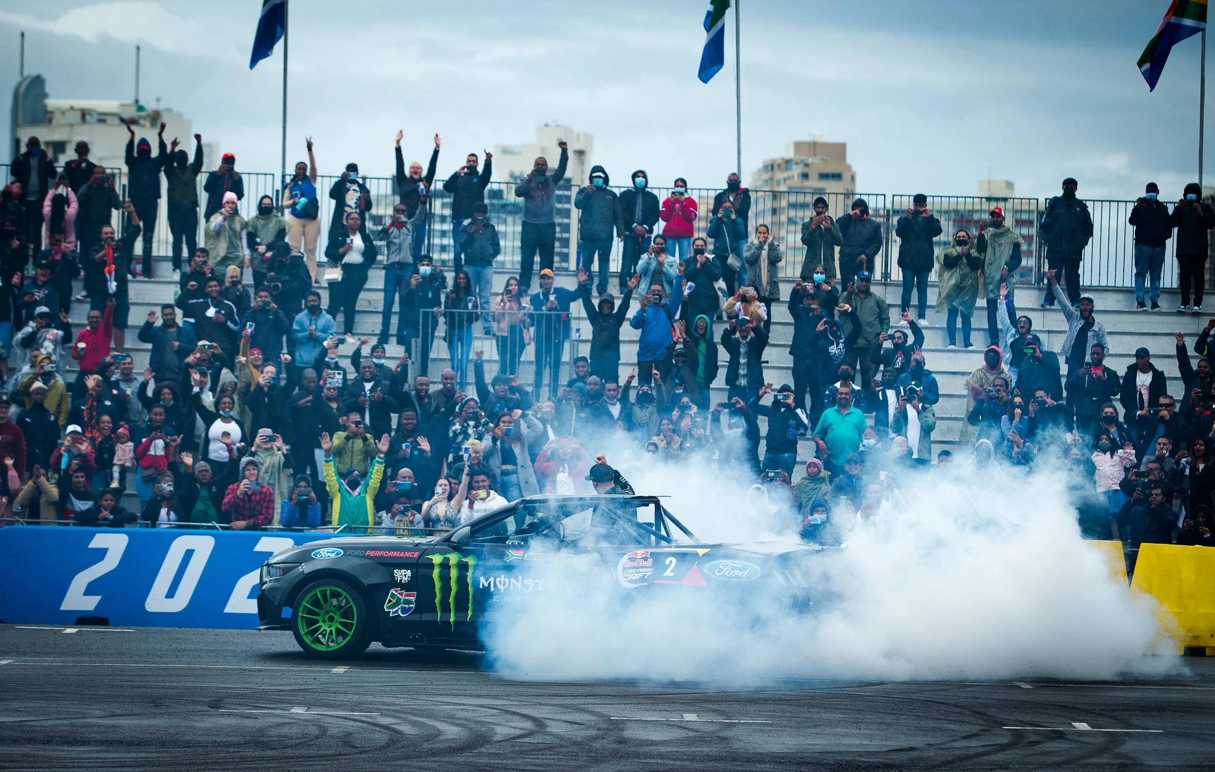 A car performing a burnout during a racing event with a crowd of spectators cheering and taking photos in the background.