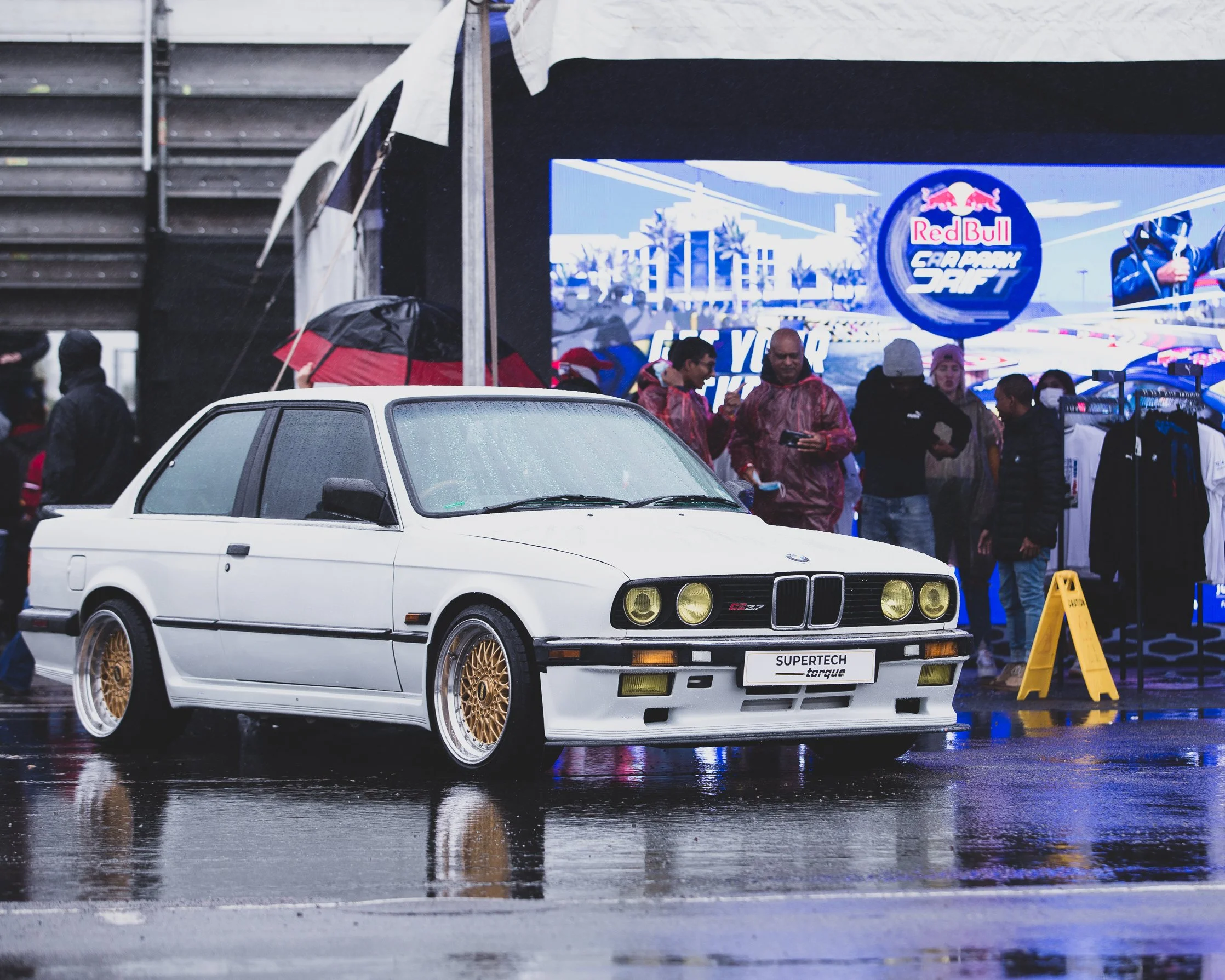 A white vintage BMW E30 M3 car with gold wheels parked on a wet surface during a rainy event, with people and a large screen in the background displaying Red Bull branding and the word 'Carpark Drift'.