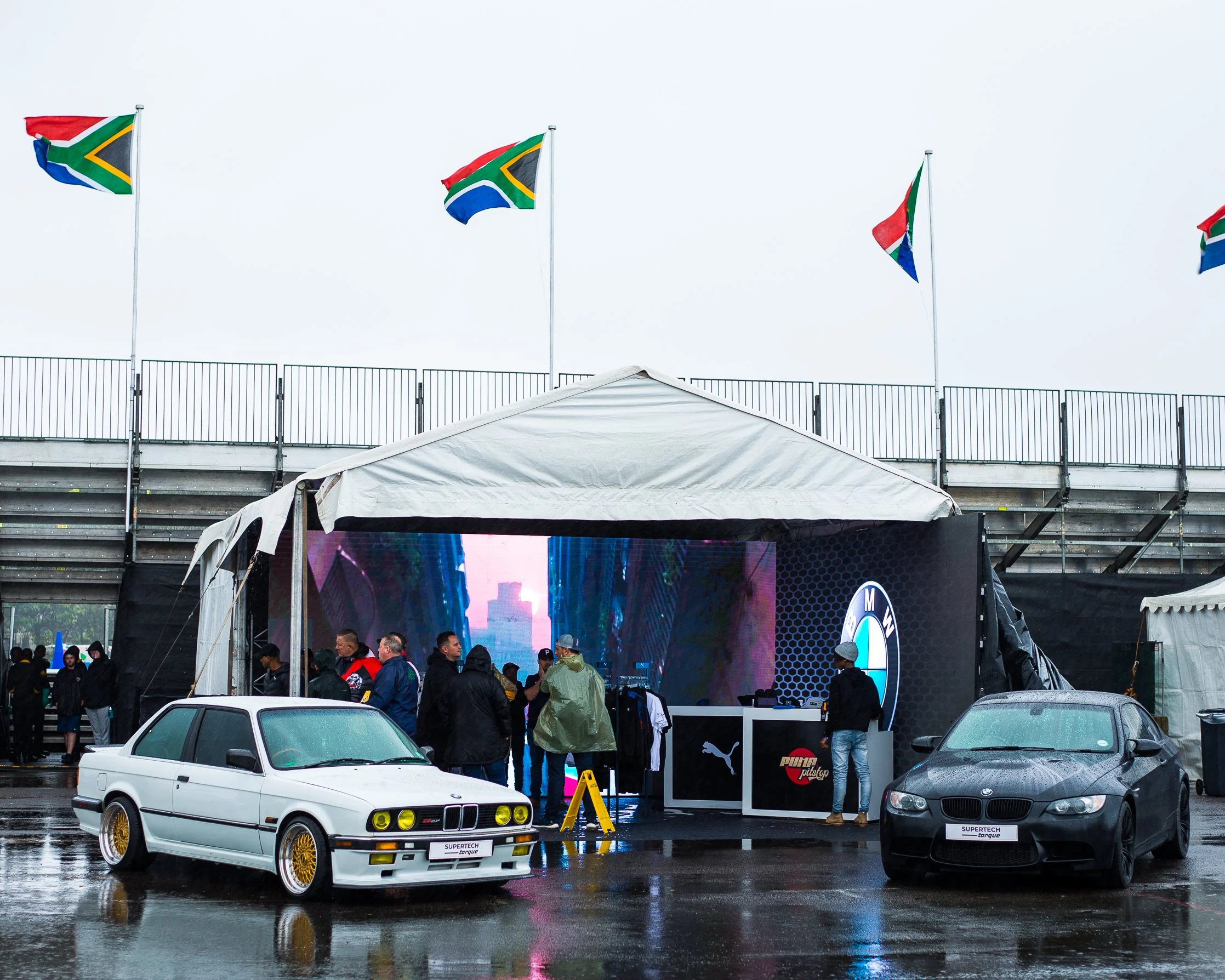 A BMW car and a black BMW car parked in front of a tent at an outdoor event, with flags flying on poles in the background and a digital screen displaying a BMW logo.