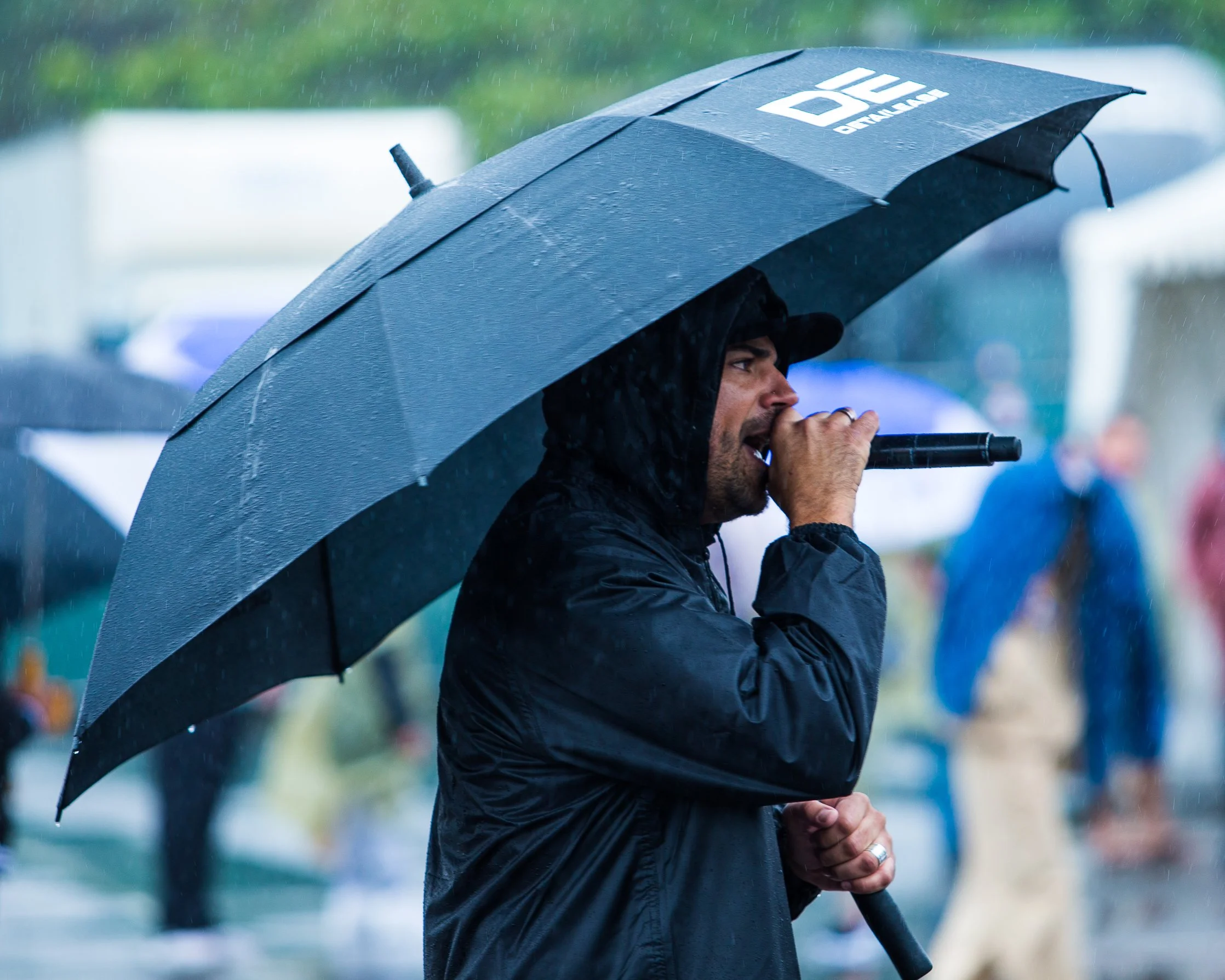 Man holding a microphone under a large black umbrella during rain at an outdoor event.