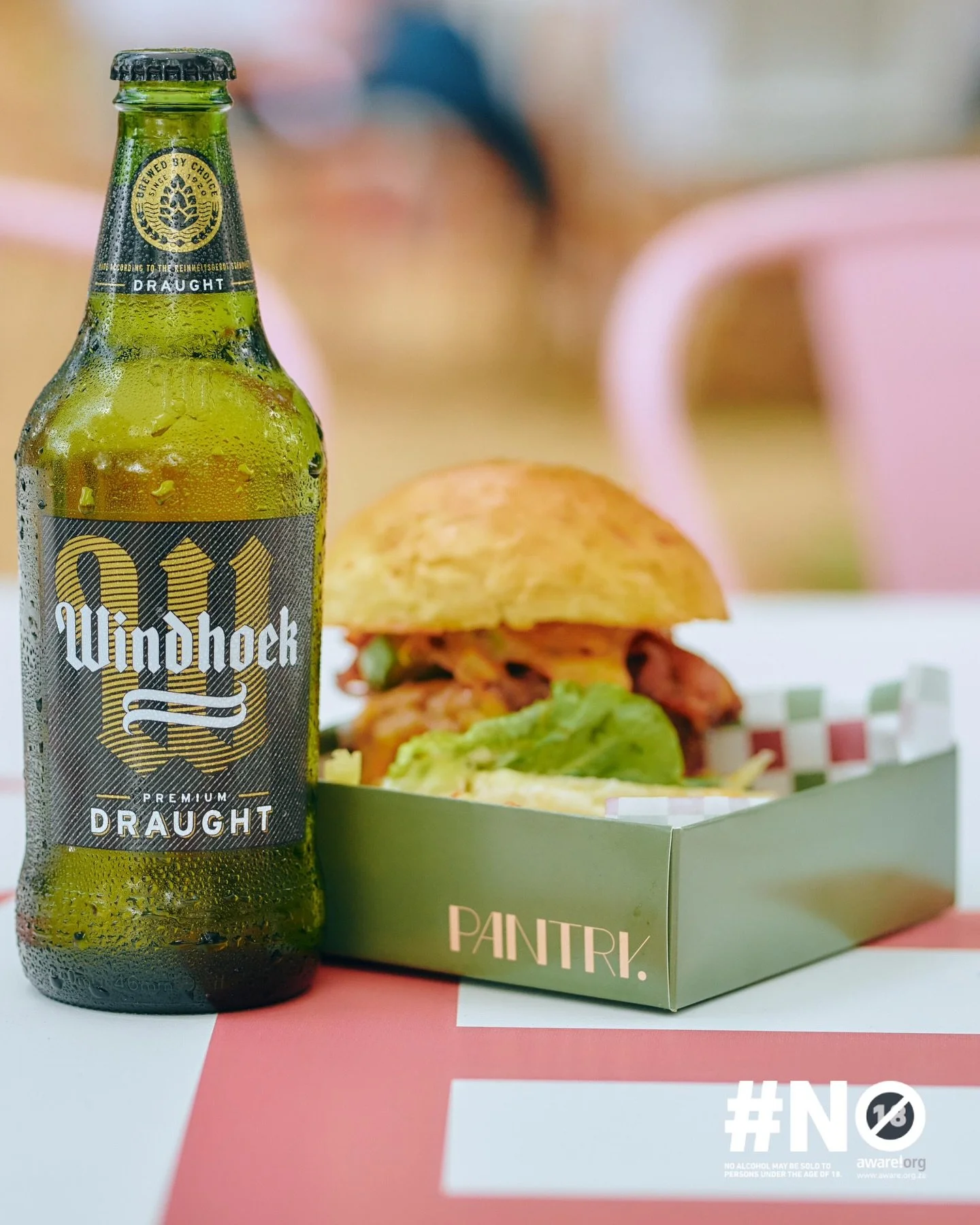 A bottle of Windhoek beer next to a sandwich with lettuce in a green box labeled 'PANTRI' on a pink and white table