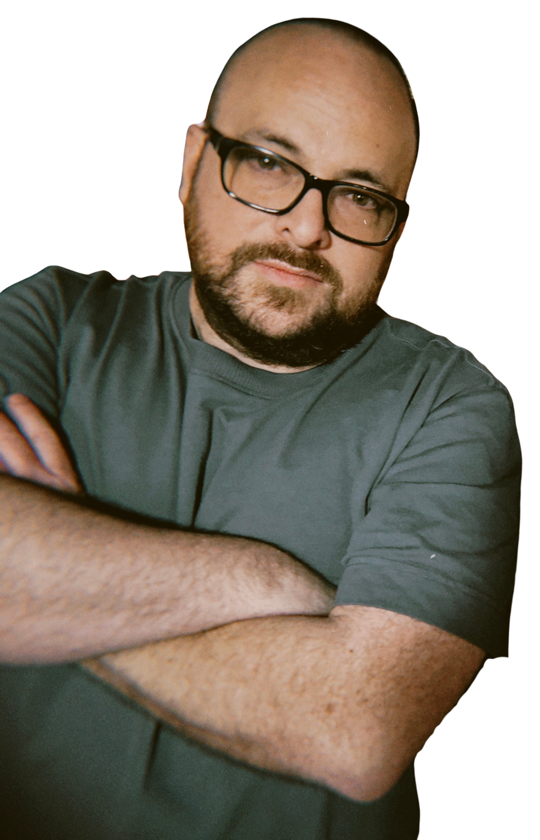 A man with glasses and a beard, wearing a gray t-shirt, has arms crossed and looks at the camera against a black background.