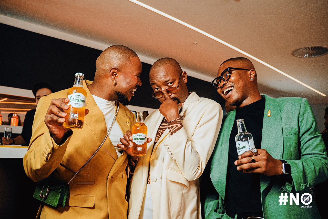 Three men dressed in colorful suits are smiling and laughing at a social gathering, holding bottles of Savanna beer.