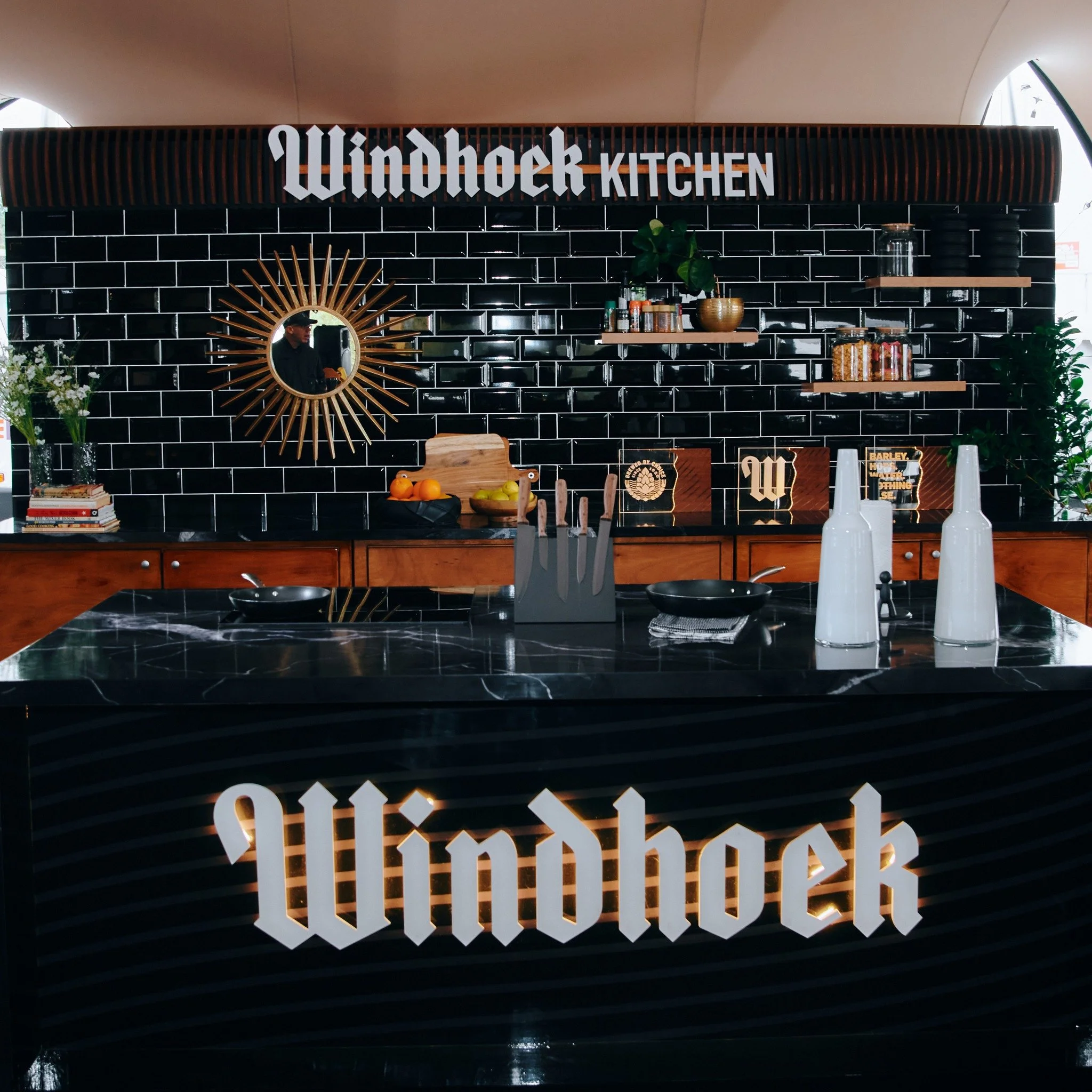 Modern kitchen with black subway tile backsplash, wooden shelves, and branded signage for Windhoek Kitchen. Features a black marble countertop, white decorative bottles, and kitchen utensils.