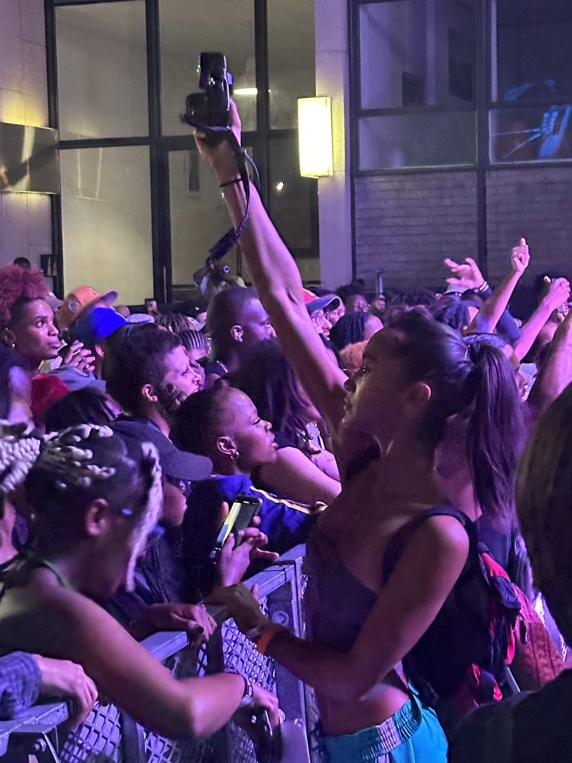 A crowd of concert-goers at a music event, with a woman in the foreground raising her arm and a person holding a camera above her. The venue has large windows and colorful lighting.