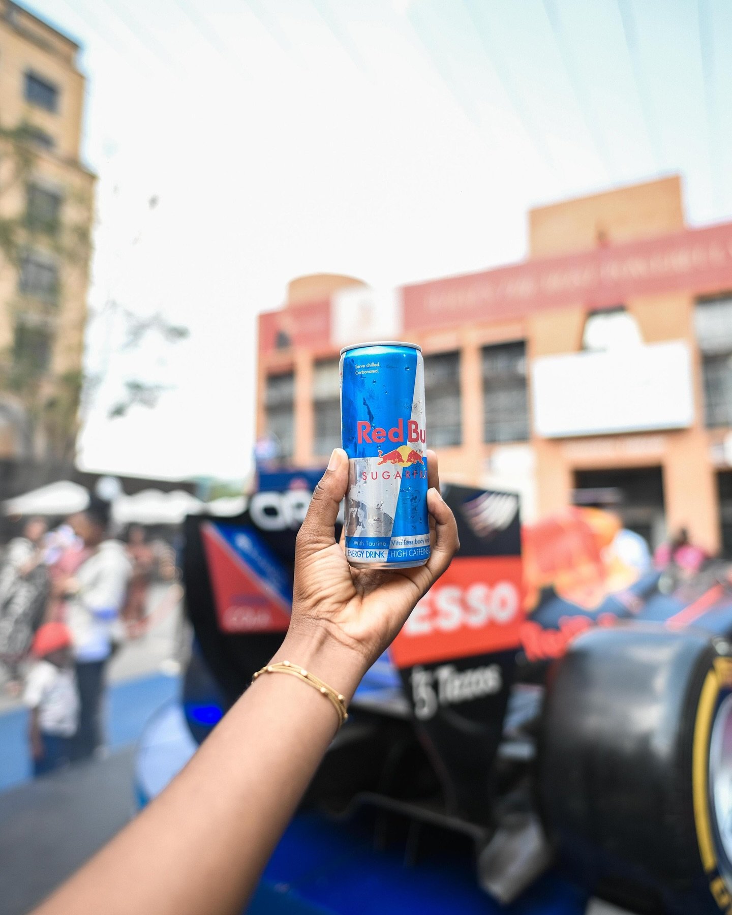 A hand holding a can of Red Bull energy drink with a blurred outdoor event in the background, including people and a racing car.