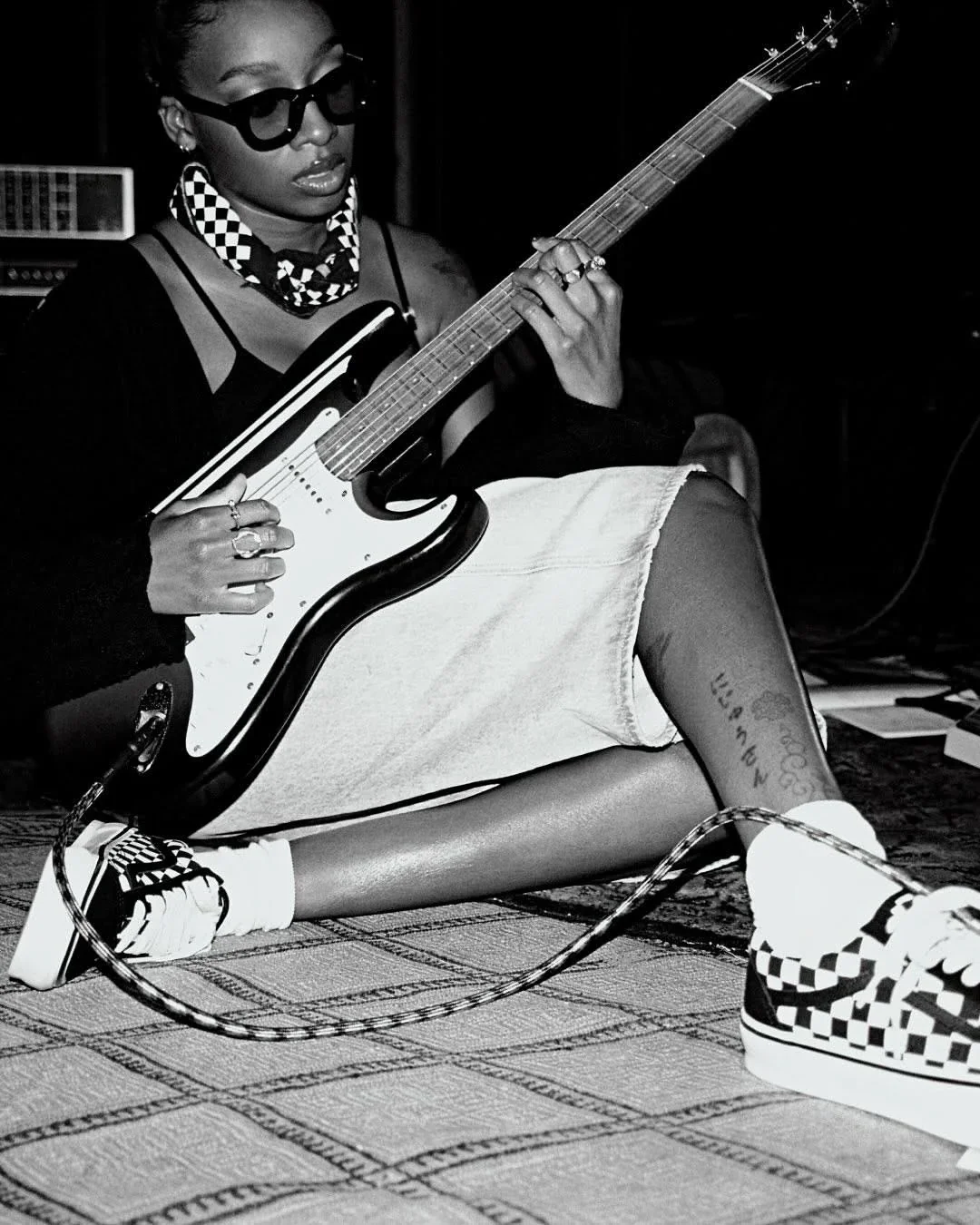 A woman sitting on the floor playing an electric guitar, wearing sunglasses, checkered shoes, and a patterned bandana, with a tattoo visible on her calf.