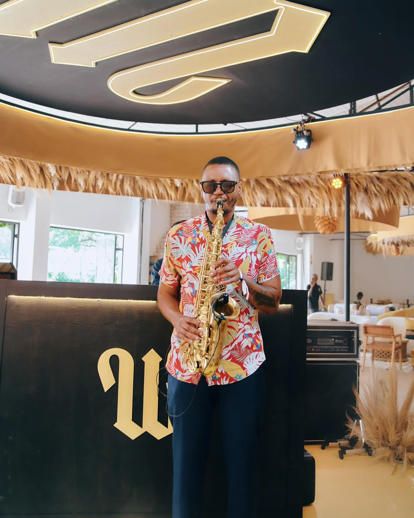 A man wearing a colorful tropical shirt and sunglasses playing a saxophone indoors at a lively event.