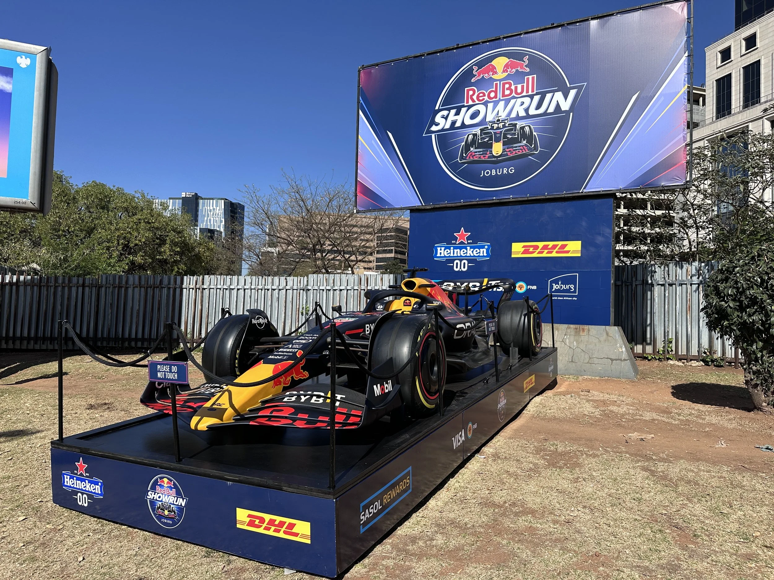 A Formula 1 race car displayed outdoors on a platform with sponsorship logos in front of a large billboard advertising the Red Bull Showrun event in Johannesburg.