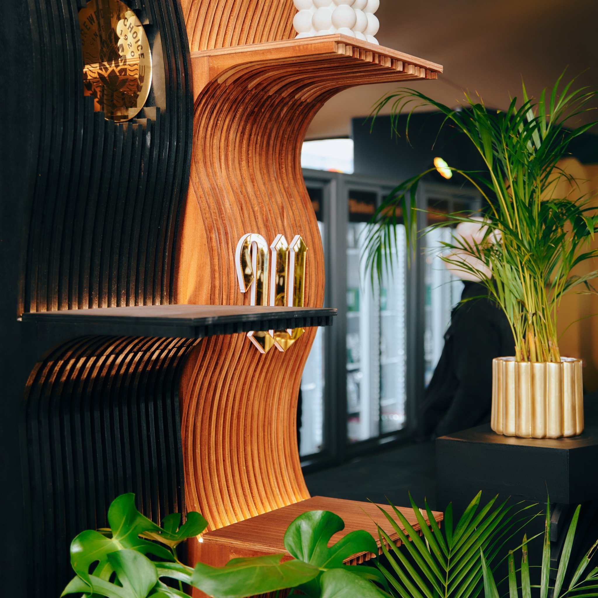 Interior of a modern cafe with a decorative wooden and black slatted shelf, gold decorative letters and a potted plant with long green leaves.