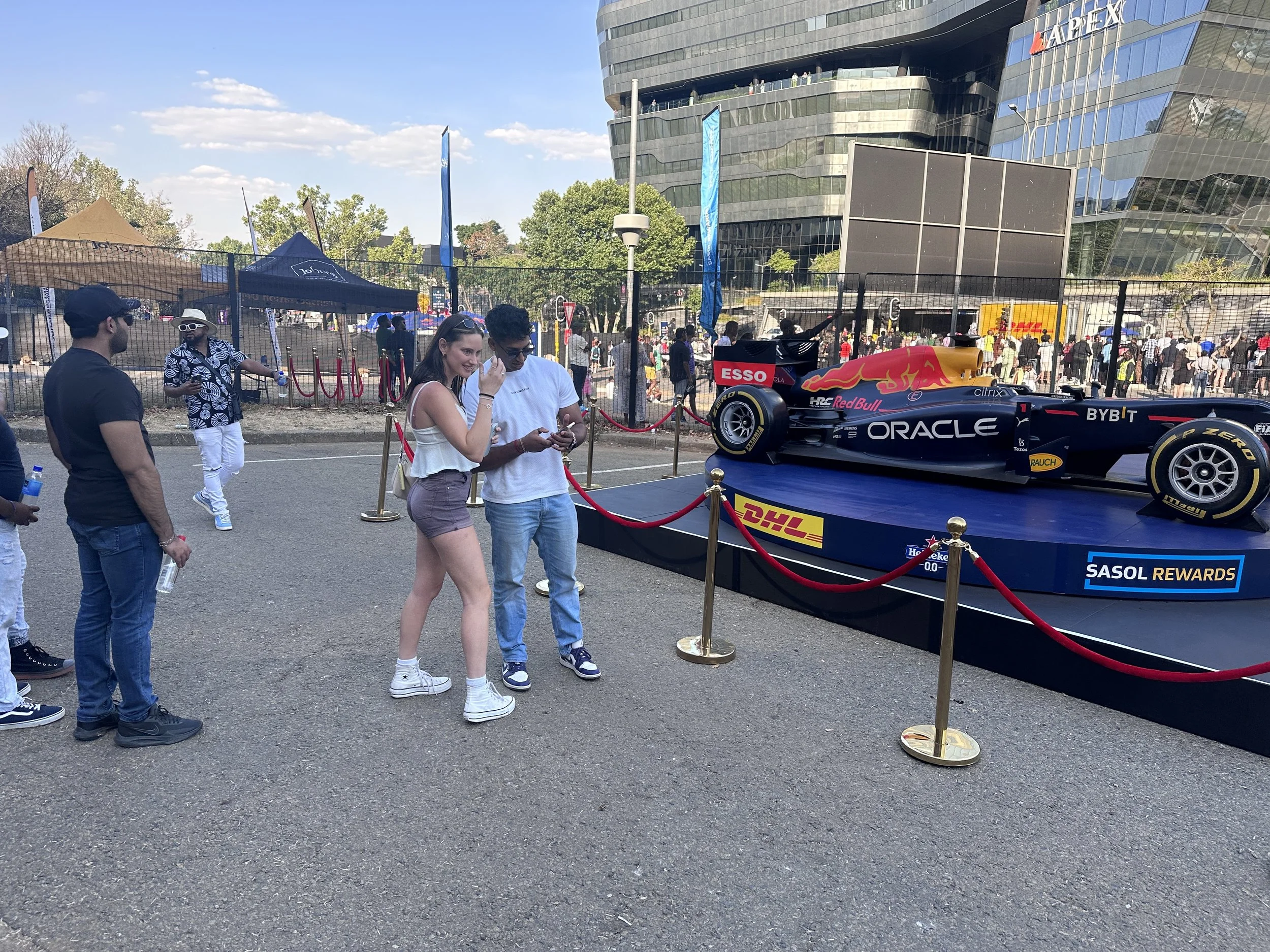 People standing outdoors near a racing car display on a raised platform, with modern buildings and tents in the background.