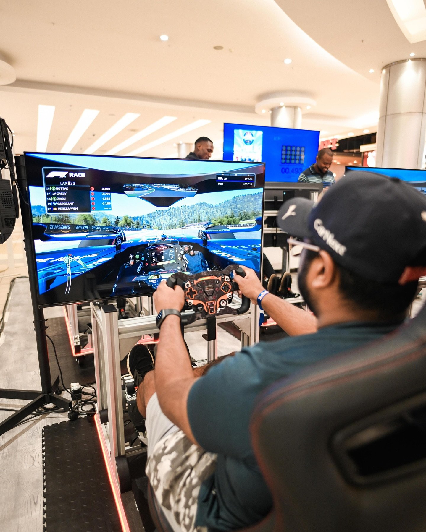 A man wearing a cap and glasses sitting in a racing simulator, holding a steering wheel controller, watching a racing video game on a large screen in a mall or indoor space.