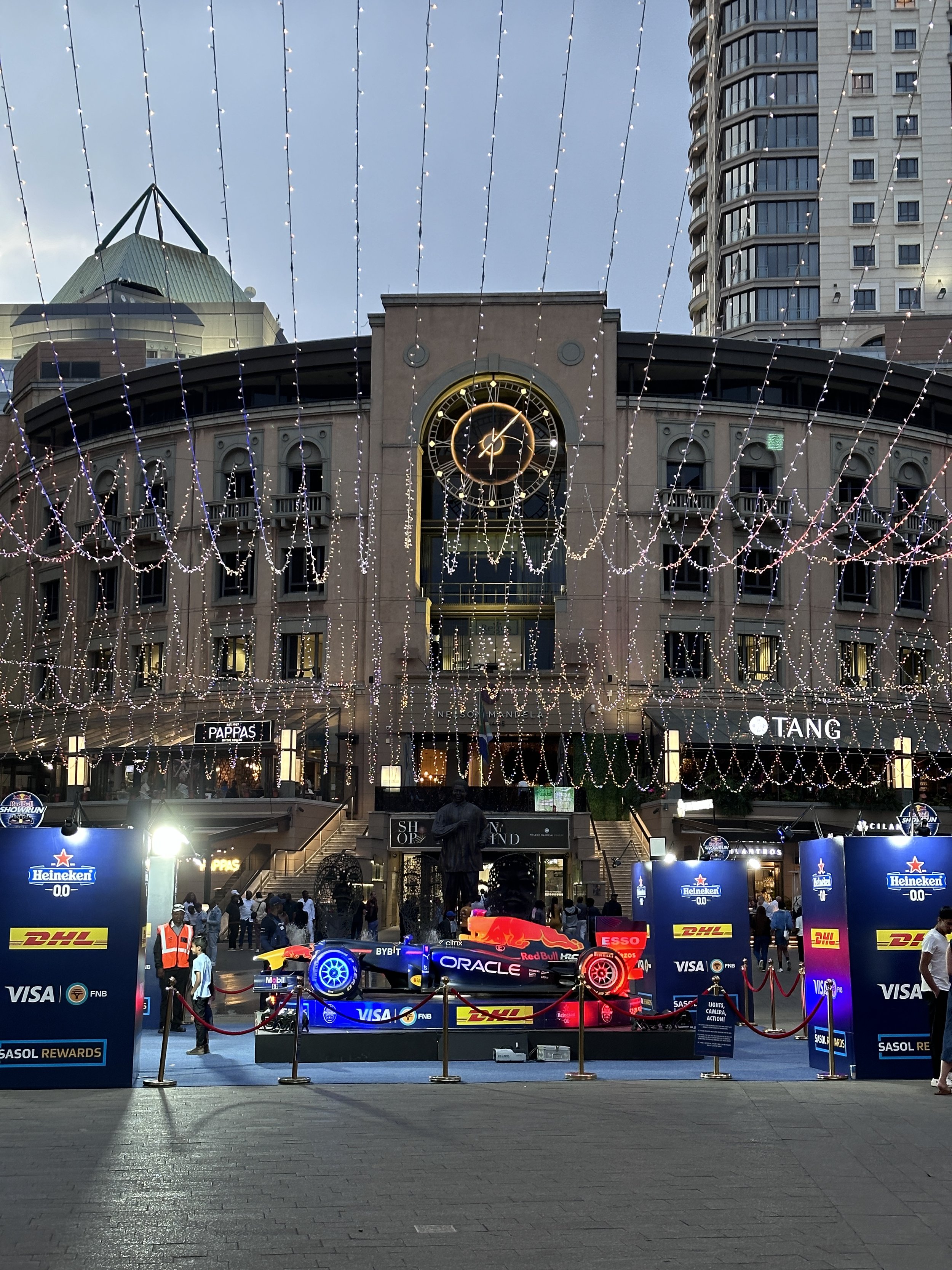 A racing car on display with sponsor logos, surrounded by barriers and staff, in front of a building decorated with string lights and a large clock, in an urban setting.