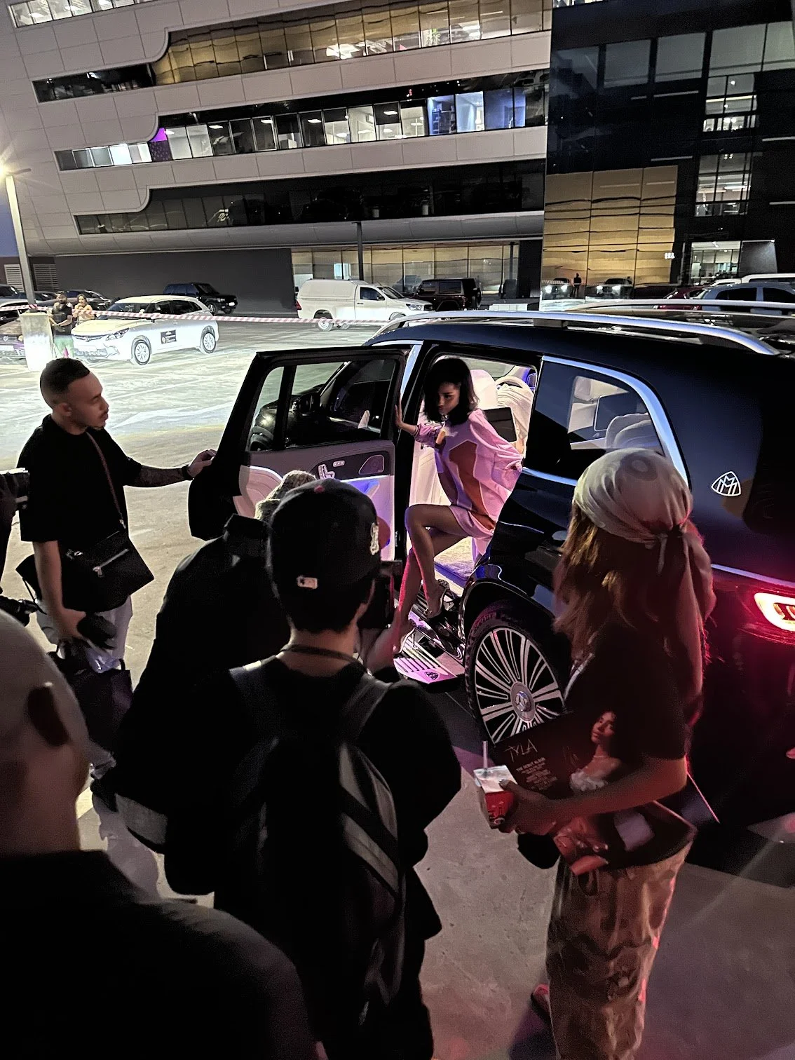 A woman in a pink dress sitting in a black luxury car, with several people around her, some taking photos, outside a modern building at night.