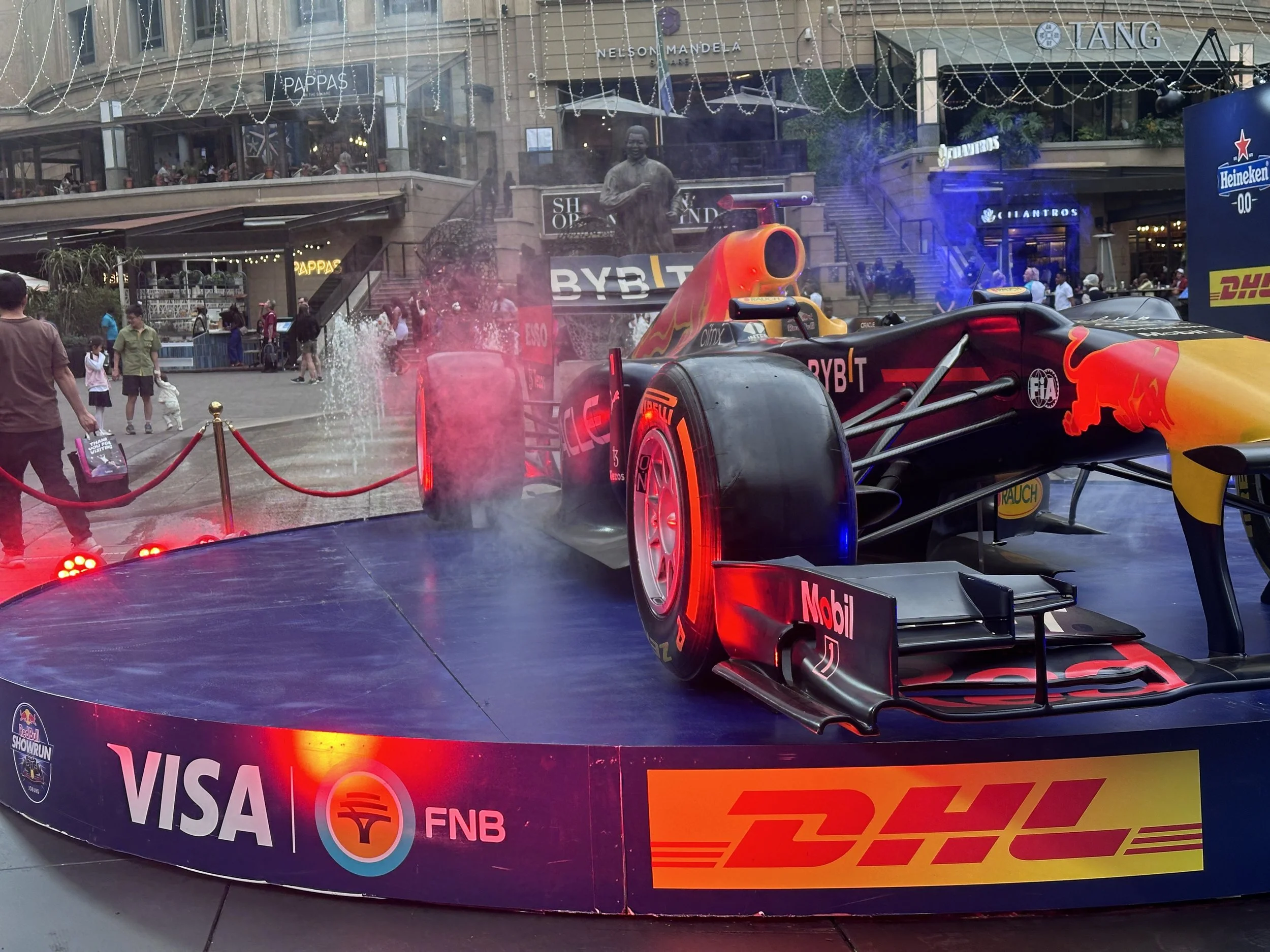 Red Bull Racing Formula 1 car displayed on a platform at an event, with the Red Bull logo, sponsors, and branding visible, surrounded by a crowd and city buildings in the background.