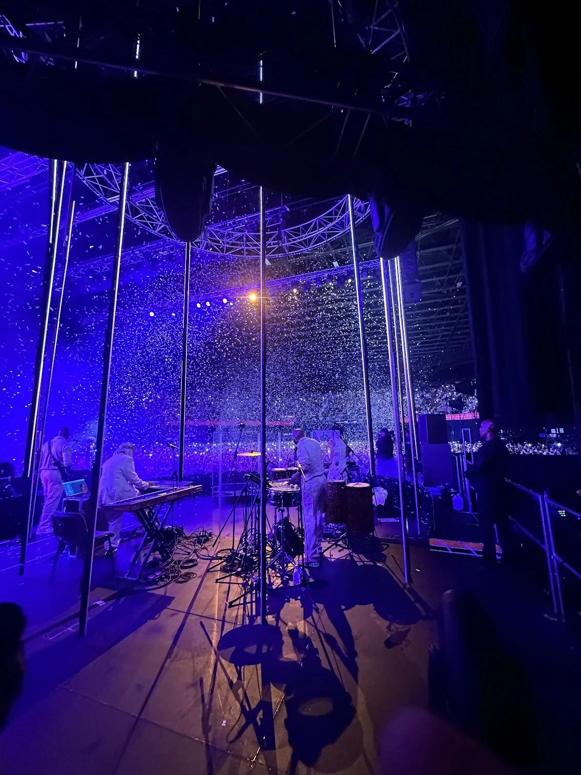 A music performance on stage with illuminated poles, confetti or snow, and musicians playing instruments in a dark setting with purple and blue lighting.