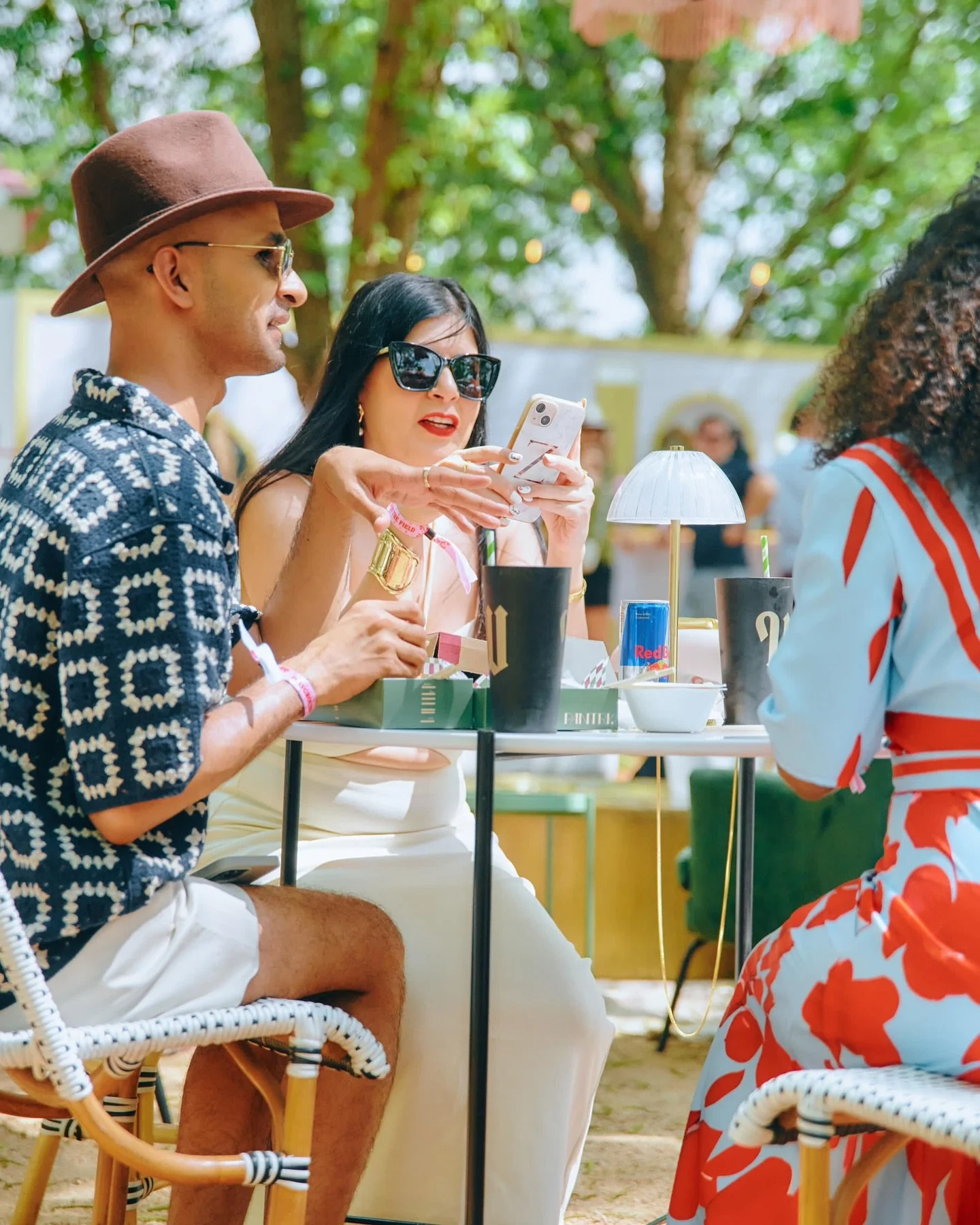 Three people sitting at a table outdoors during daytime, two women and one man, with trees and other people in the background. The woman in the center is looking at her phone, wearing large sunglasses and red lipstick, the man on the left is wearing 