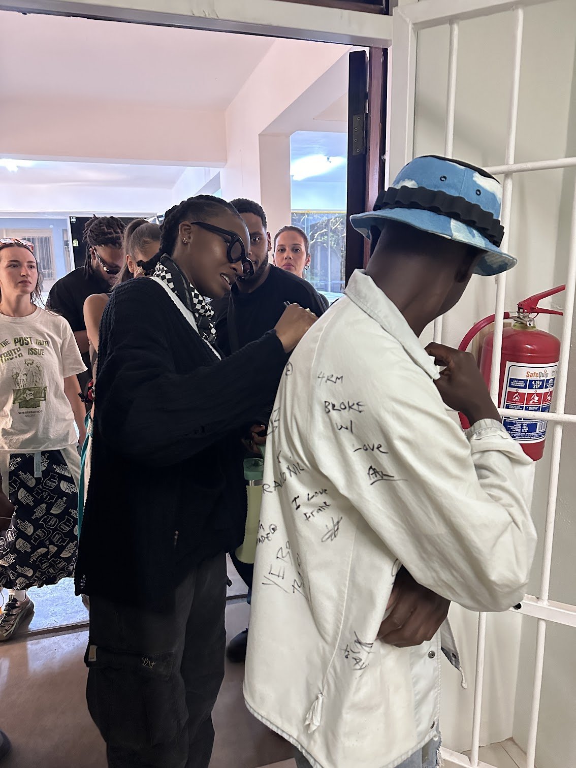 A man is applying a patch to another man's white jacket, which has various signatures and writings on it, while a group of people watch.