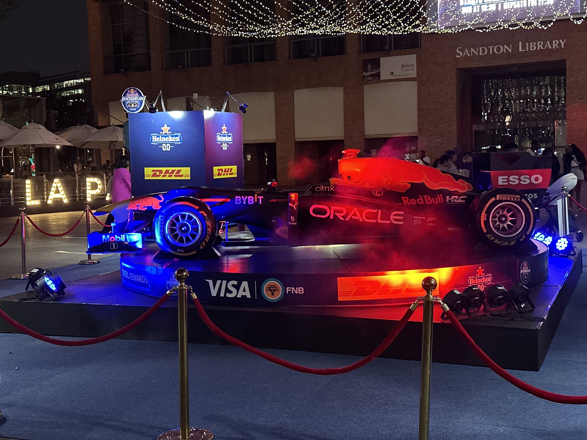 A Formula 1 race car on display outdoors at night, illuminated with red and blue lights, surrounded by a red velvet rope barrier, with sponsors' logos including Oracle, Red Bull, Heineken, and Visa visible on the car and platform.