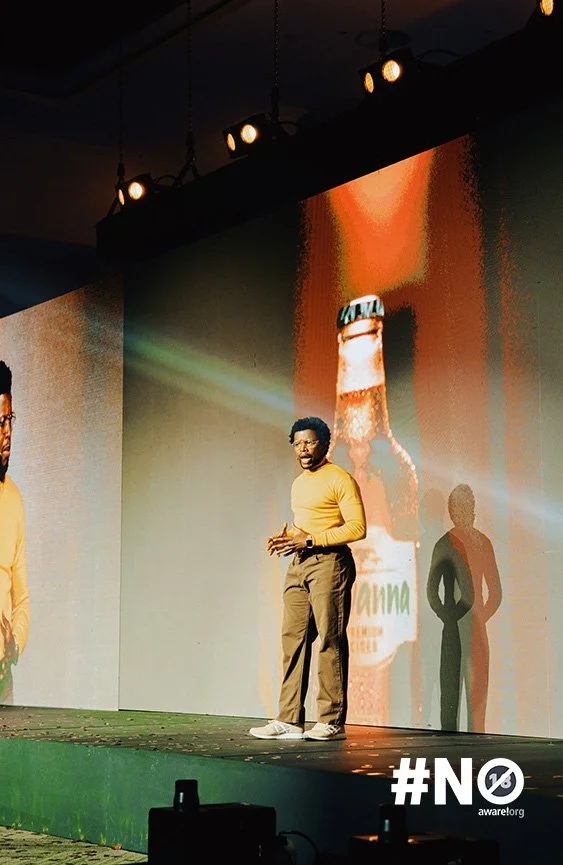 Man speaking on stage in front of a large screen with beer bottle image, wearing a yellow sweater, brown pants, and white shoes, with stage lighting and a hashtag #NO at the bottom right.