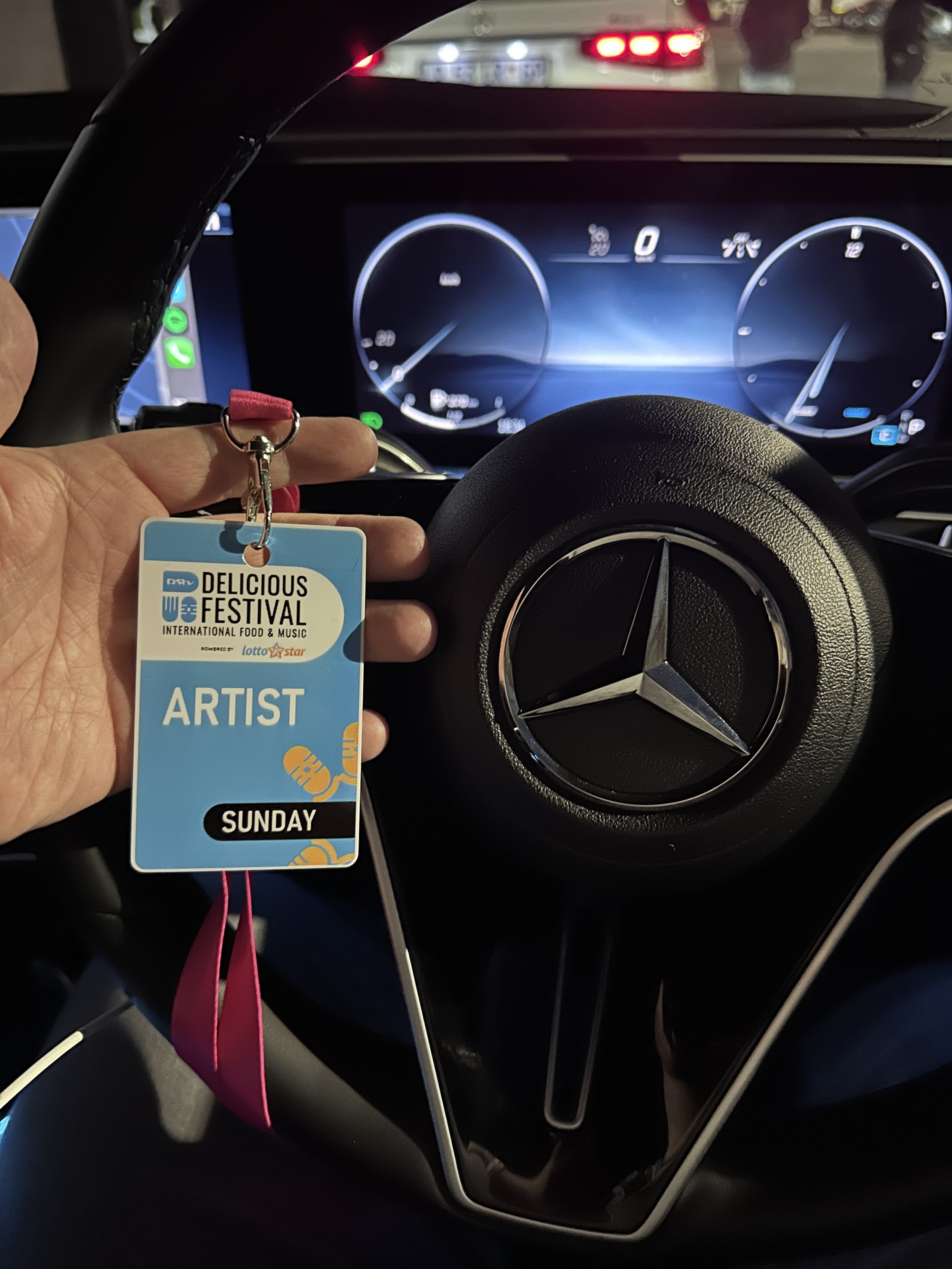 Photo of a person's hand holding a festival badge in a Mercedes-Benz car, with a digital dashboard in the background.