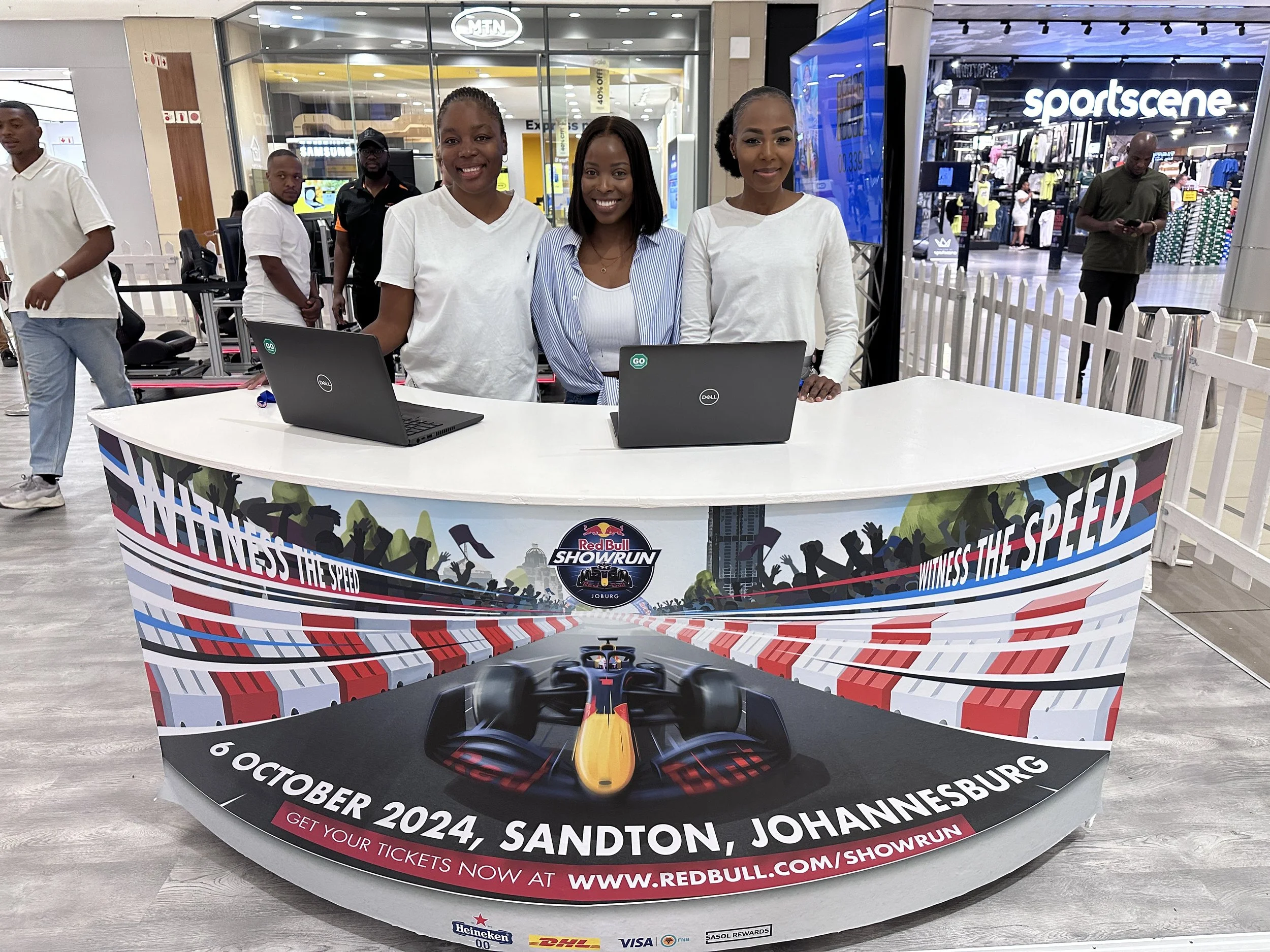 Three women standing behind a promotional table for the Red Bull Showrun event, with two laptops on the table; the event is scheduled for October 6, 2024, in Sandton, Johannesburg, with details to get tickets at the website listed.