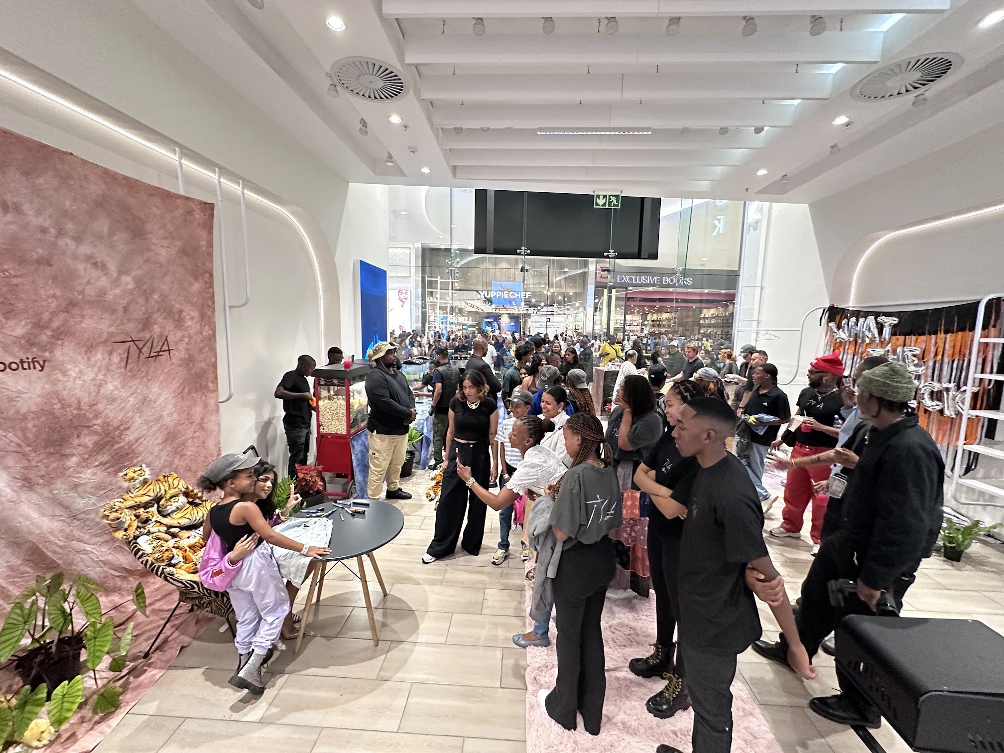 Crowd of people gathered at an indoor event in a mall, with tables, decorations, and a popcorn stand, as children and adults socialize.