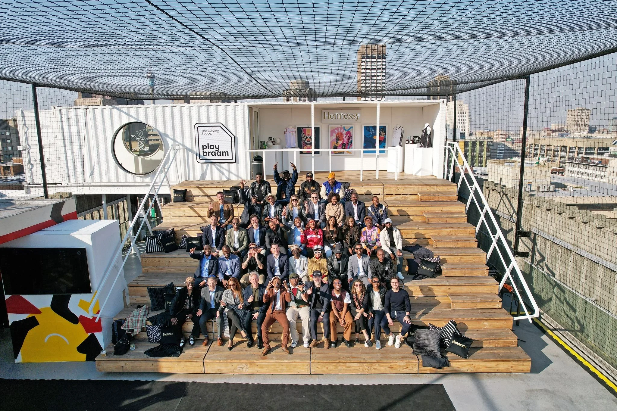 Group of people sitting and standing on a multi-level wooden deck on a rooftop with city buildings in the background, during a sunny day.