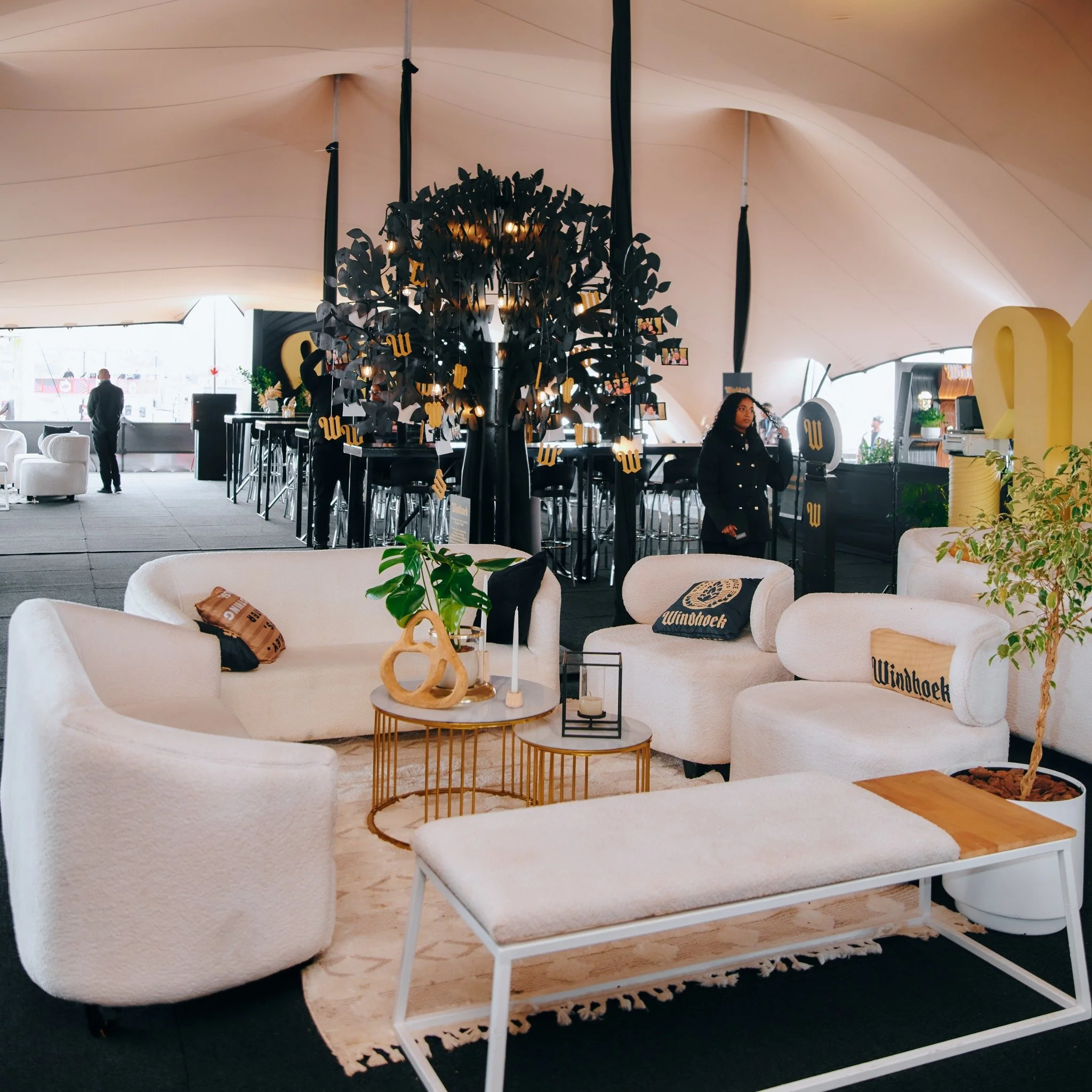 Indoor lounge with white sofas, decorative pillows, a round coffee table, potted plants, and a large black tree-like structure with hanging golden accents, under a curved ceiling.