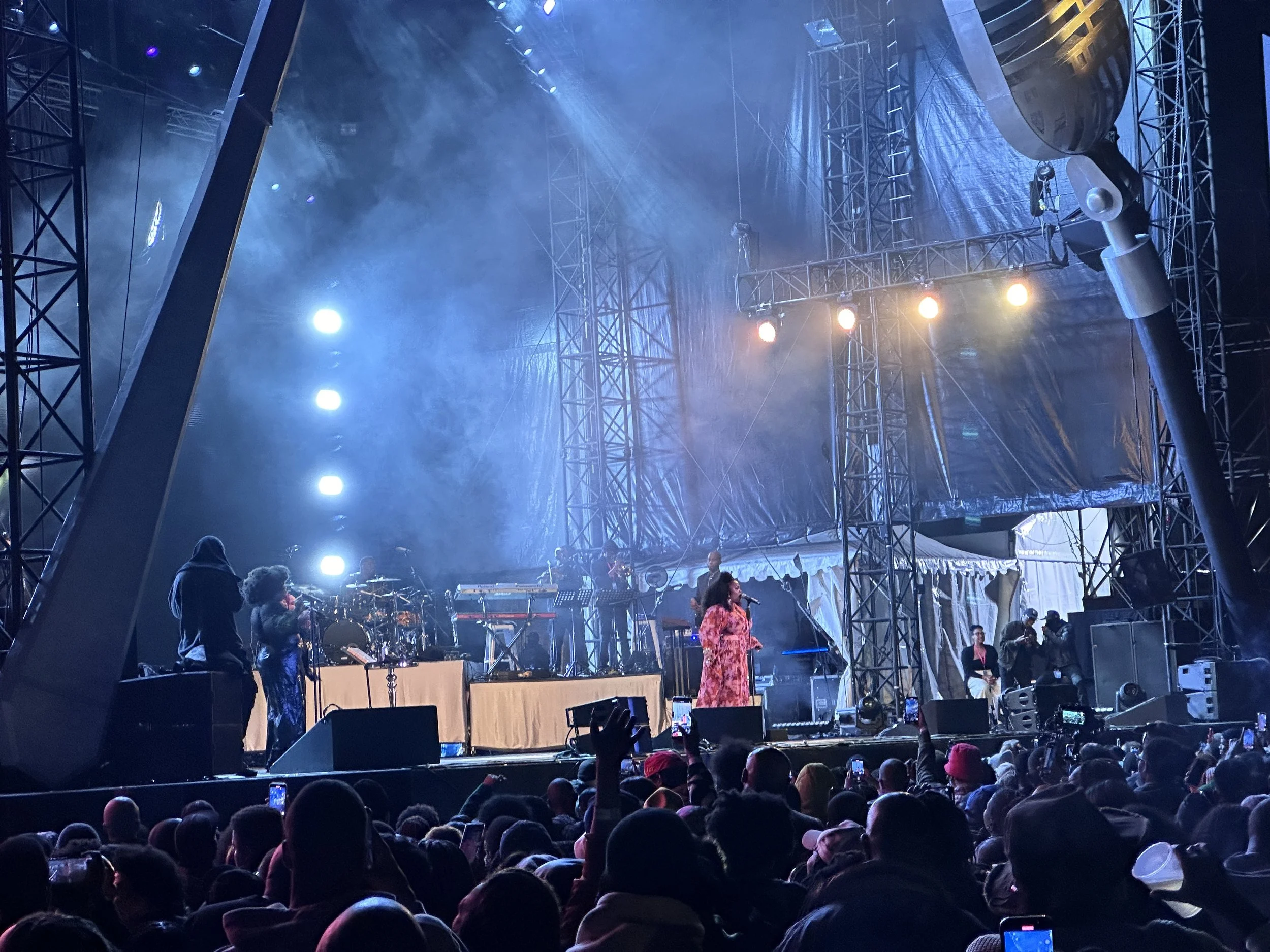 A concert stage with performers and bright stage lights, audience in the foreground, and a large structure supporting the stage.