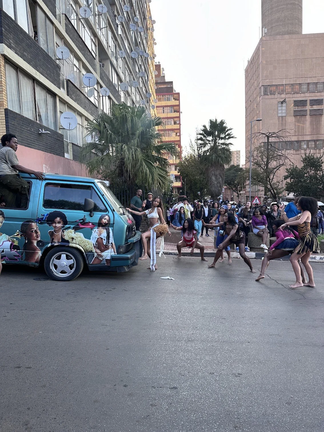 People dancing on the street next to a decorated vehicle with large posters of women, in an urban area with buildings, palm trees, and a crowd watching.
