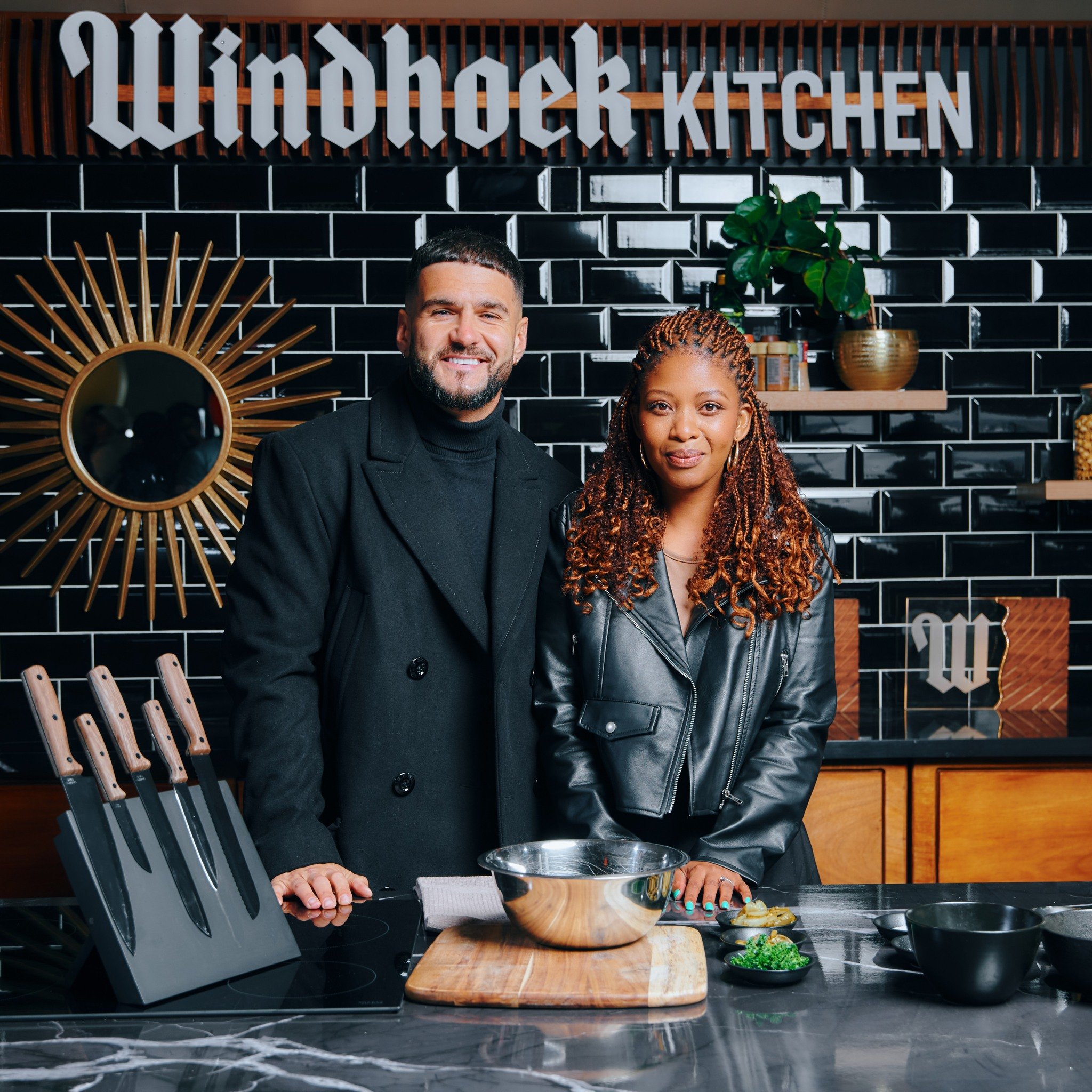 Two people standing in a modern kitchen with black tiled walls, a wooden shelf with plants, and a sign that reads 'Windhoek Kitchen'. They are smiling, and there are knives on a block, a metal mixing bowl, and various ingredients on the counter.