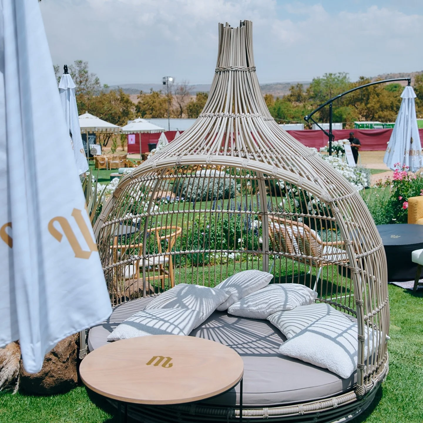 Wicker outdoor sunbed with cushions and pillows, shaped like a dome, situated within a garden area with umbrellas and flowers, under partly cloudy sky.