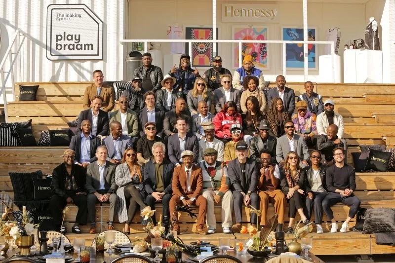 Group photo of diverse people sitting and standing on wooden-tiered seating at a Hennessy event with modern decor and branding.