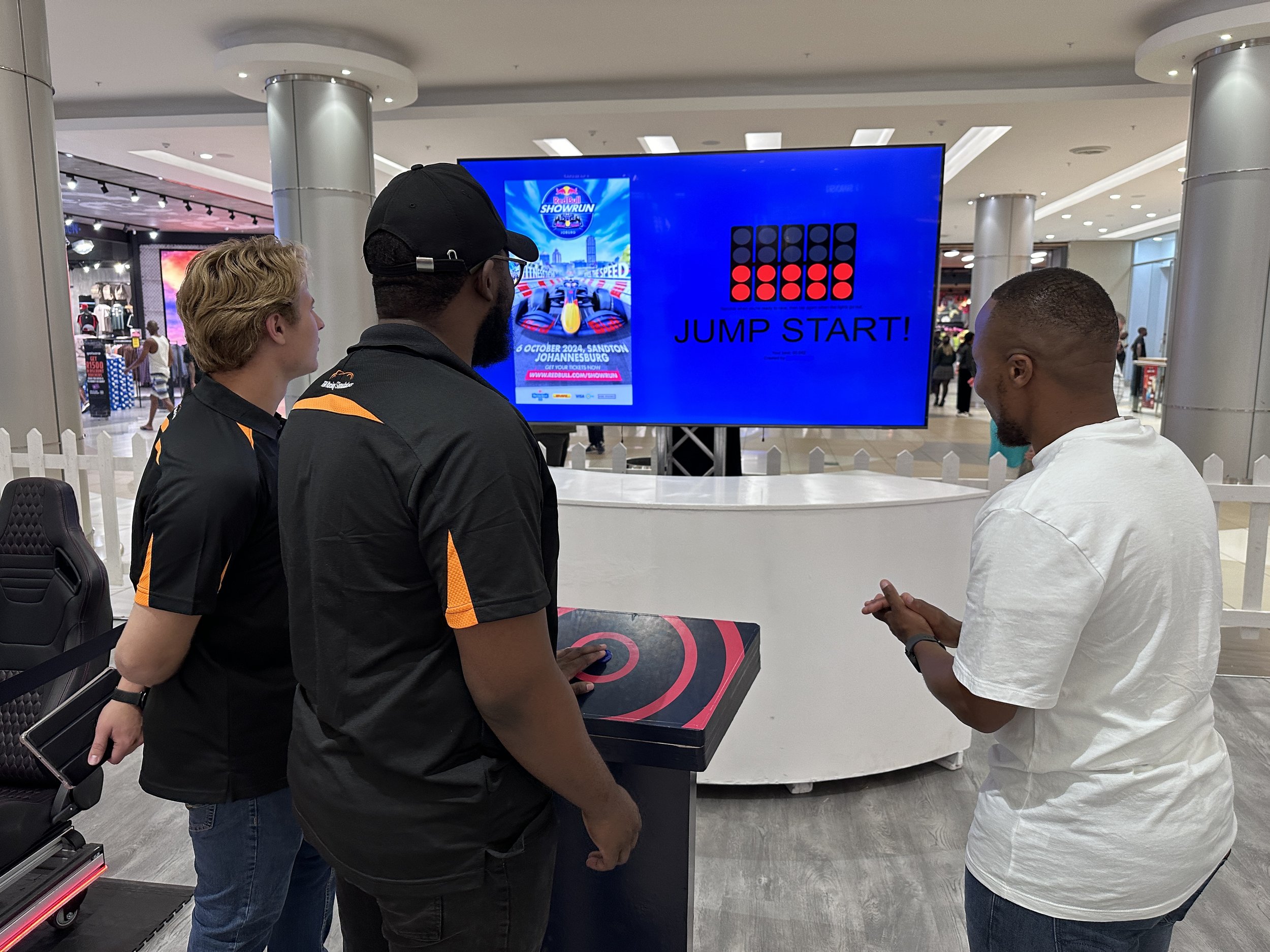 Three men are playing a game at an arcade in a shopping mall, with a large digital screen behind them displaying the words "Jump Start!" and an event poster.