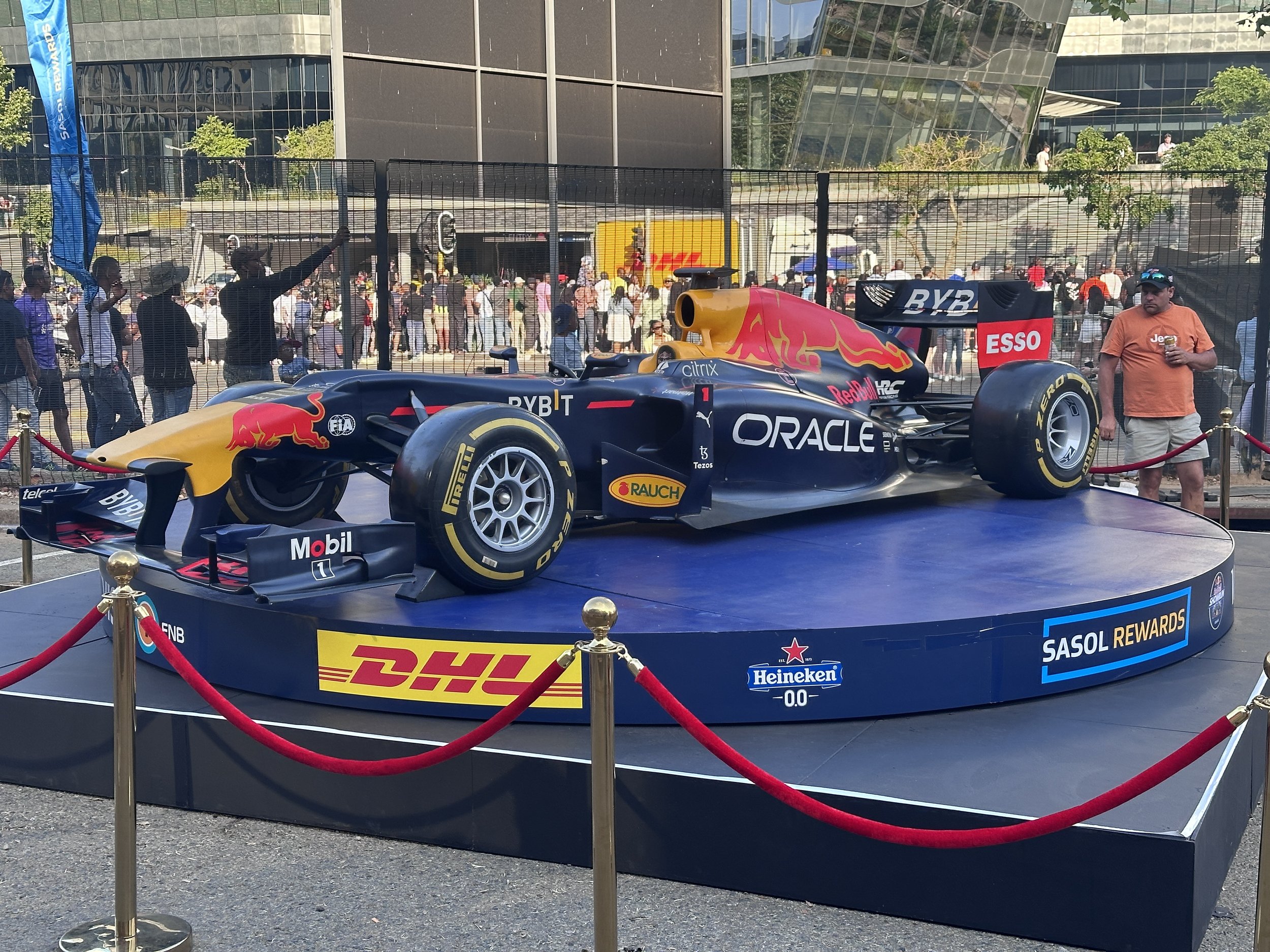 A red bull Racing F1 car displayed on a circular platform with banners from DHL, SASOL REWARDS, and Heineken. The car features logos from Oracle, Mobil, Pirelli, and other sponsors. The display is surrounded by red ropes, and a crowd of people is see