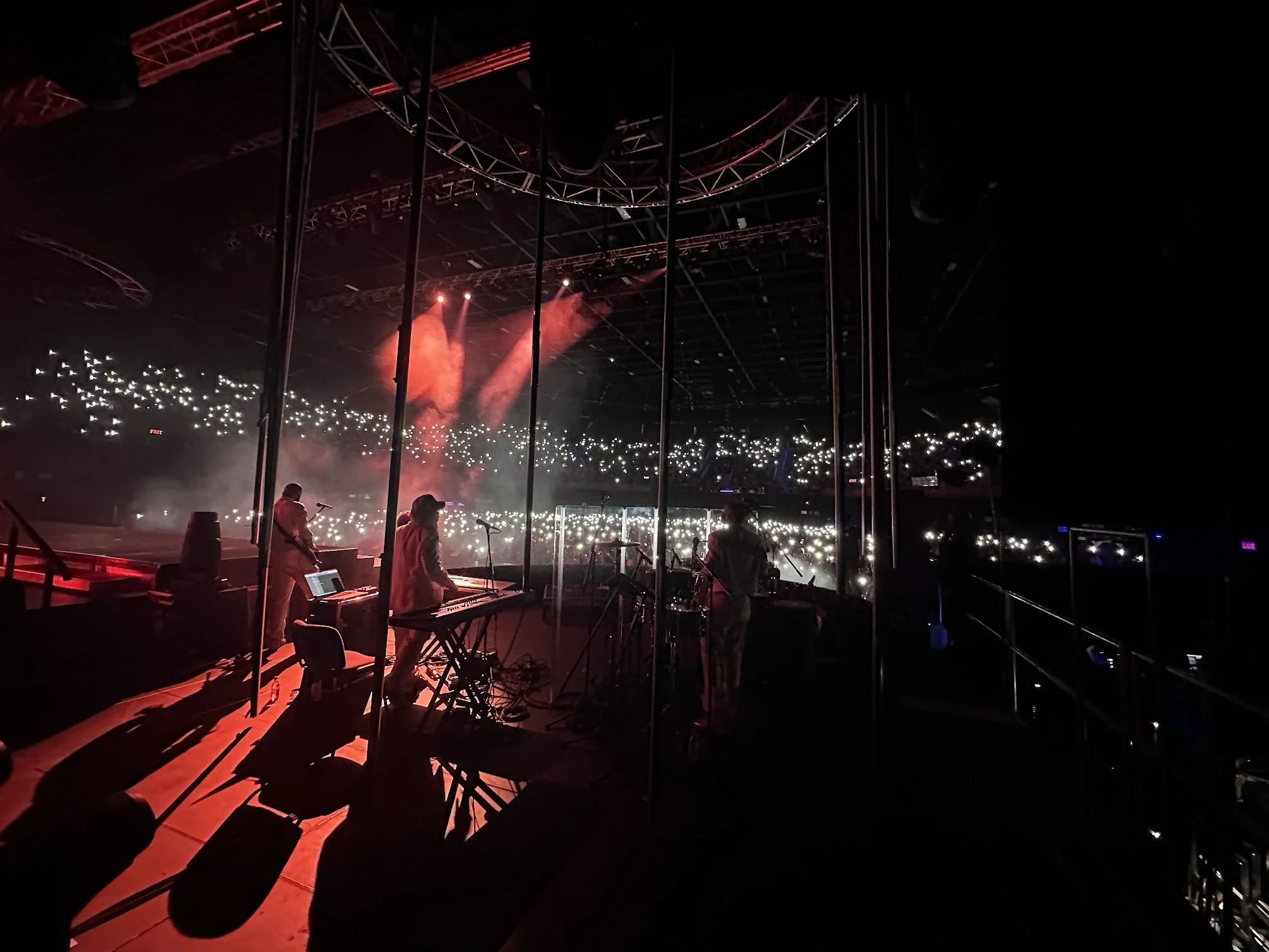 Band performing on stage with red lighting and smoke effects, audience holding up lights in a dark concert venue.